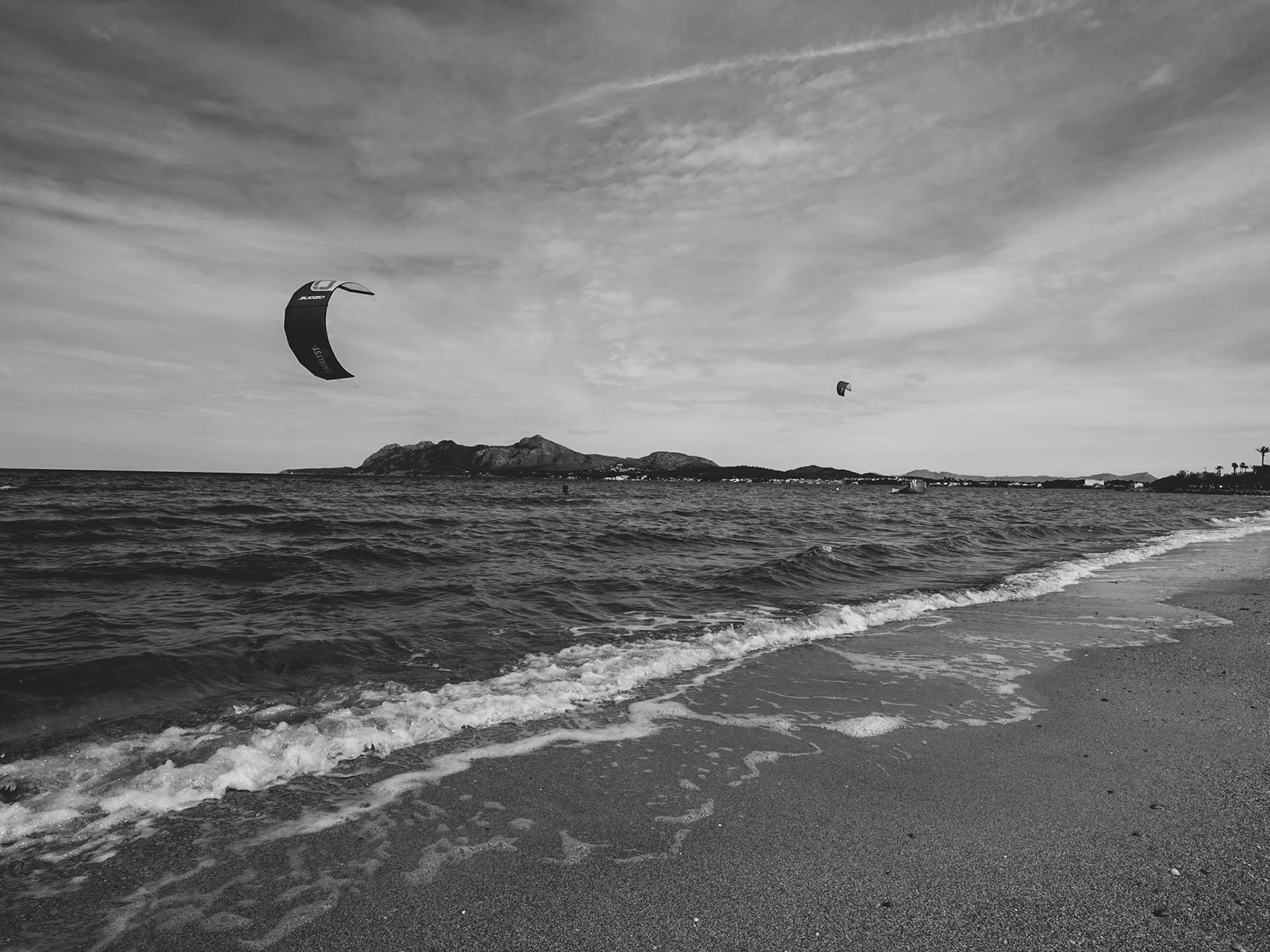 in the bay of pollença, a sanctuary for wind and kite surfers, the monochrome palette speaks in contrasts. here, the wind is an artist, sculpting waves, dictating the dance of the kites above. the northeastern coast of mallorca whispers of adventure with its generous breezes and waves, paired with a flat, long sandy shore offering a canvas of possibilities. the kites, distant yet distinct, are like thoughts set free in the vastness of the sky—black on white, simplicity on complexity. the shoreline, with each gentle caress of the surf, tells stories of encounters between land and sea, of nature's playful moods, and of spirits eager to ride the edge of the wind's melody.