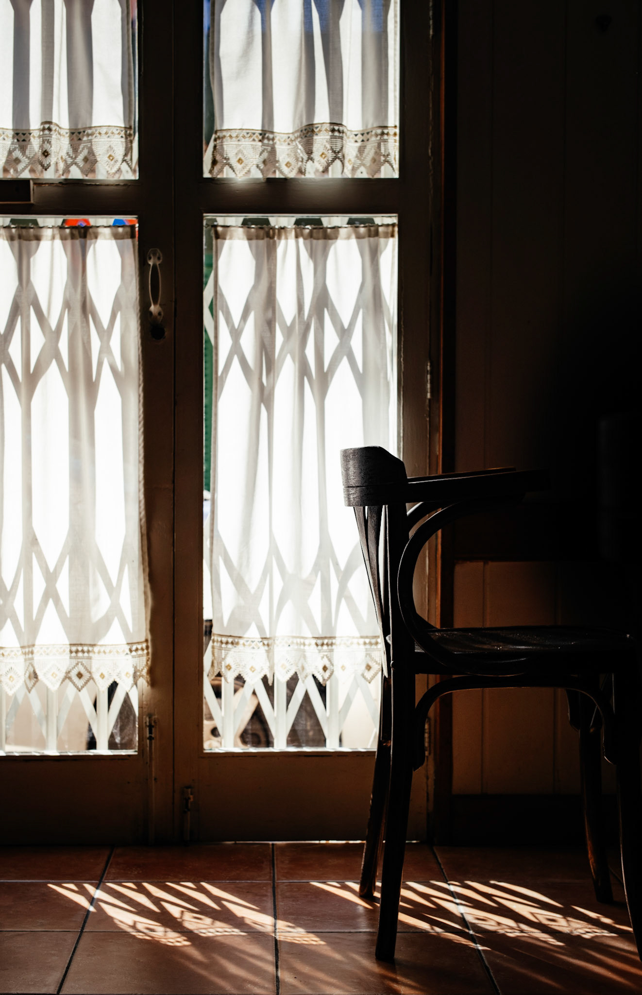 in the subdued light of "el viejo y el mar" in the pere garau district, a lone chair sits by a window. sunlight filters through lace curtains, casting intricate shadows on the tiled floor. the stillness of the scene in palma de mallorca suggests a quiet moment before the day unfolds.