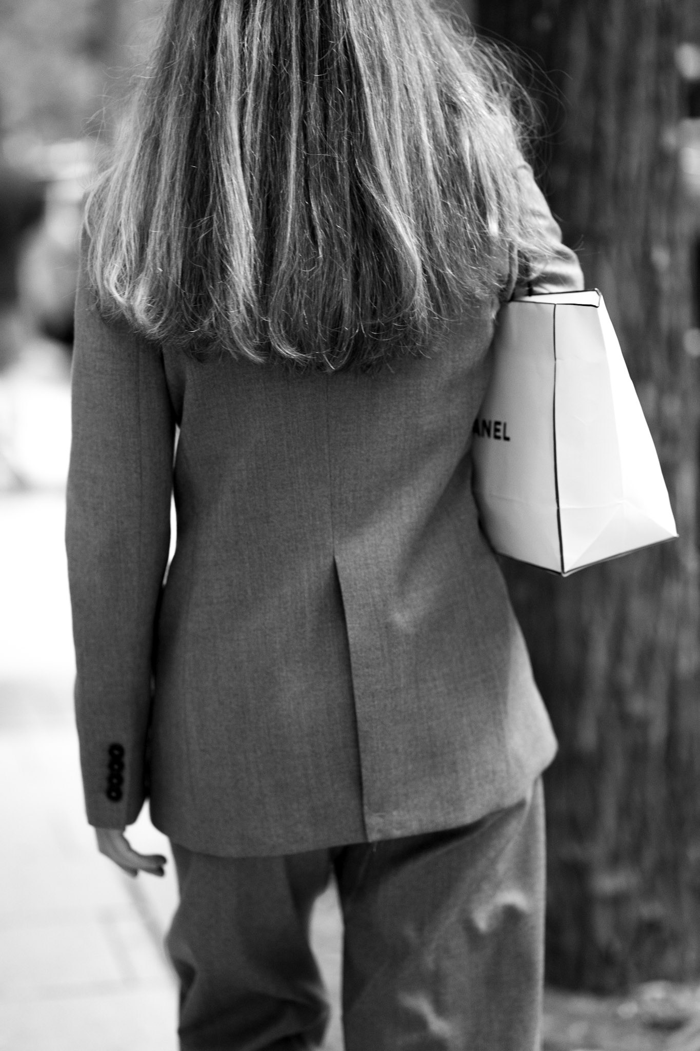 this image captures a moment of refined minimalism on the streets of madrid. with her back turned and chanel bag casually tucked under her arm, she moves through the city with effortless style. the monochrome tones highlight the simple lines of her outfit, while the textures of the blazer and bag add subtle depth. her presence is soft but striking, embodying an unspoken sophistication that blends into madrid’s urban rhythm. this shot frames the allure of quiet luxury in everyday life.
