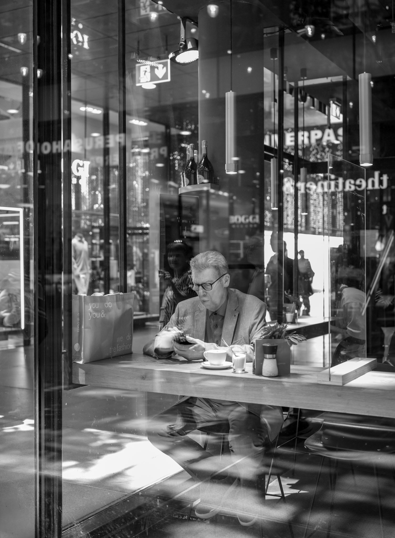 taken in the 5 höfe in munich, this photo showcases a man quietly sitting in a café, engrossed in his phone. captured through the café’s glass windows, the reflections of the surroundings create a complex and multi-layered scene. the various layers of reflections, the people walking outside, and the modern architecture create a rich visual experience. the black-and-white format enhances the contrasts and textures, giving the image a timeless atmosphere. this photo captures the stillness and contemplation amidst the hustle and bustle of urban life, inviting the viewer to explore the deeper layers of everyday life.