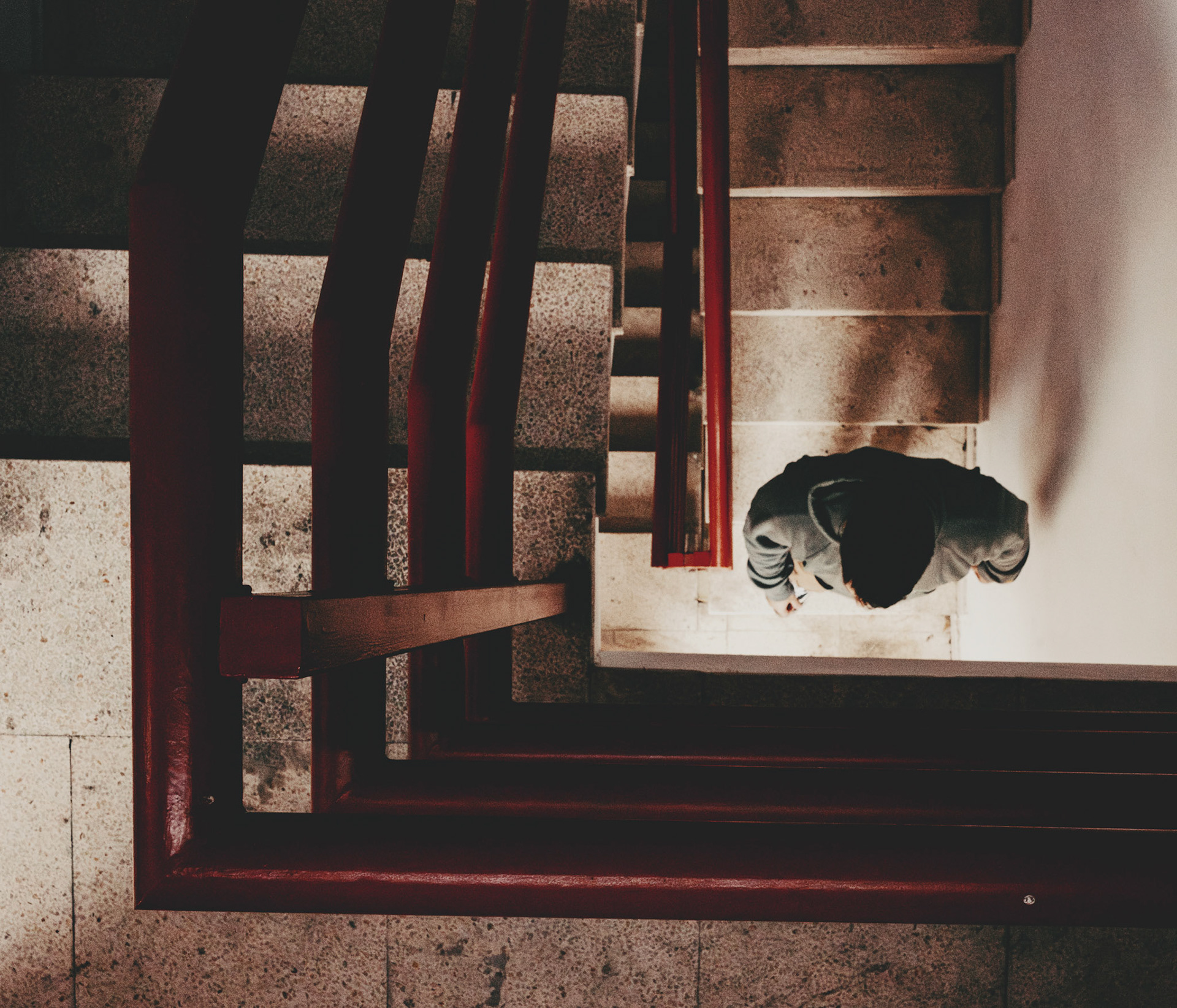 captured in a parking garage in palma de mallorca, this photograph offers a compelling perspective on the mundane act of descending stairs. the geometric lines of the red handrail frame the scene, drawing the eye towards the figure walking down, cloaked in shadow. the interplay of light and shadow, along with the angular composition, creates a dynamic visual narrative that transforms an ordinary moment into a captivating image.
