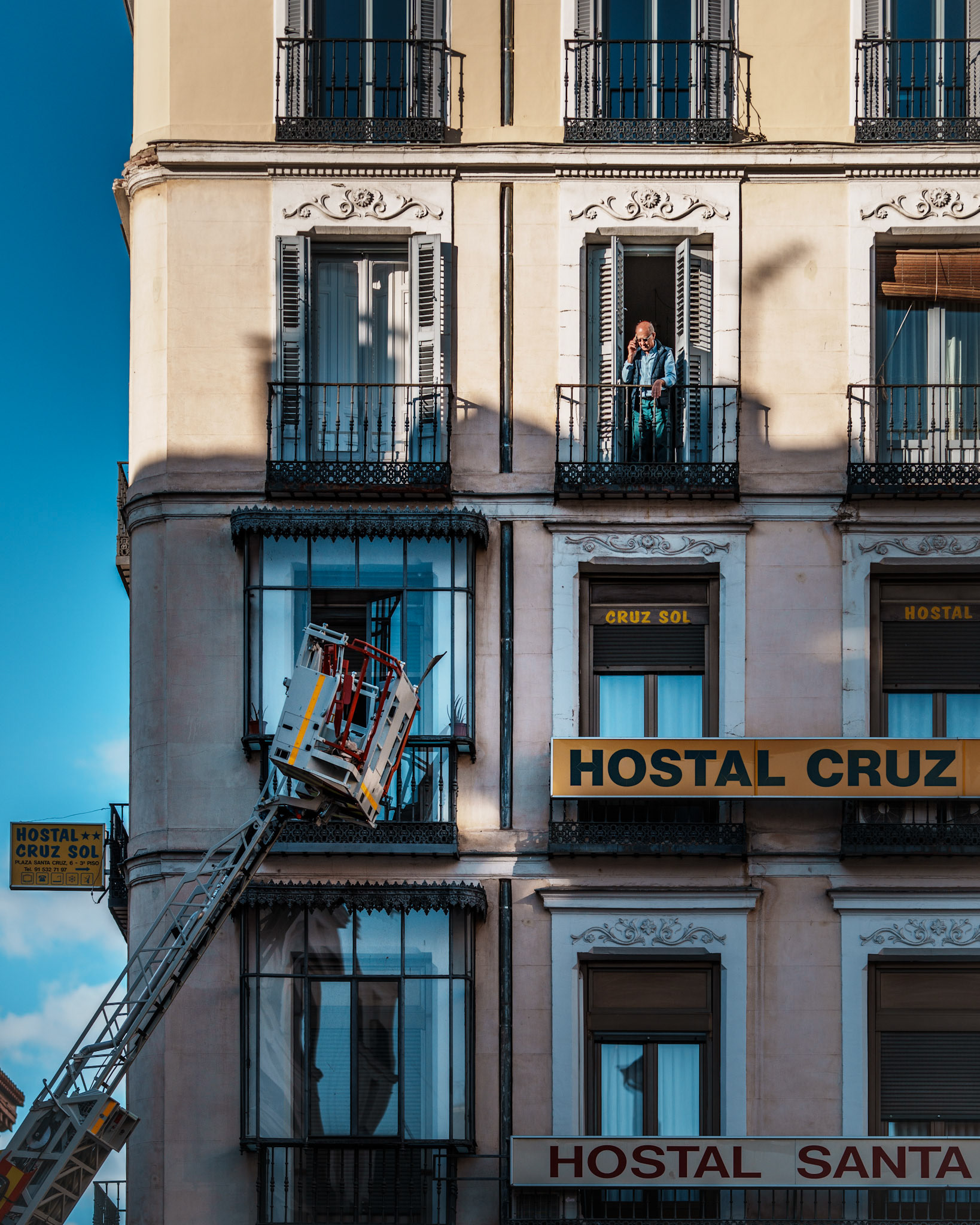 caught in a moment of pure puzzlement, this man appears to be on the phone, perhaps with the hotel reception, seeking an explanation for the unusual “external lift” that’s ascended to his balcony. the warm sunlight highlights the building’s classical details, creating a striking contrast with the modern lift awkwardly hanging below him. there’s a quiet humor in this scene, as if he’s both amused and perplexed, wondering if the hotel’s “concierge service” has been taken a bit too literally. the madrid fire department had arrived due to concerns that parts of the building’s rooftop were at risk of falling. fortunately, the man faced no fire or immediate danger, adding a touch of irony to his bemused expression.