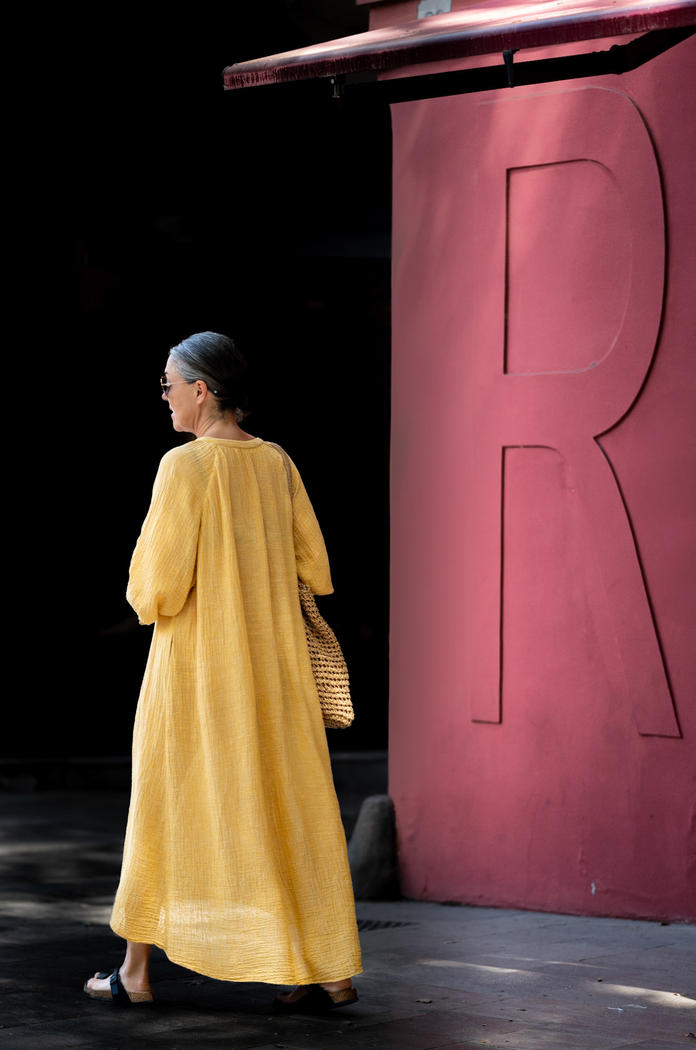 the woman in a flowing, mustard-yellow dress moves with purpose through the streets of Palma, the vivid hue of her garment contrasting starkly against the muted tones of the city. she strides confidently past the bold, red "R" emblazoned on a column, her steps echoing on the cobblestone beneath. a woven bag hangs loosely from her shoulder, hinting at a story untold, an errand incomplete, or a journey only just begun. this is a moment captured in transition — between destinations, between thoughts, between the ordinary and the extraordinary.