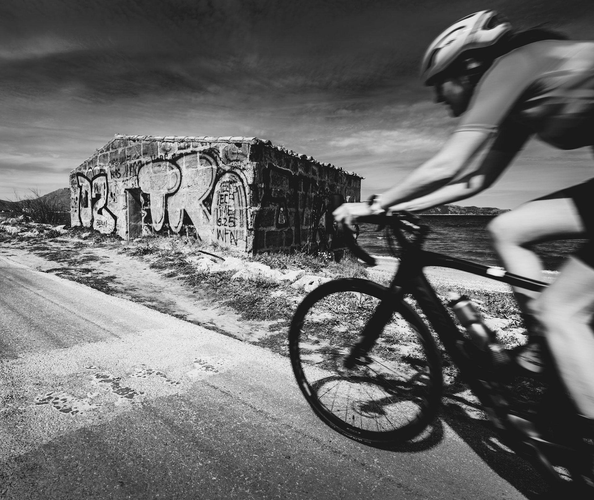 along the bay of pollença, a cyclist slices through the frame with kinetic energy, a blur against the permanence of a graffiti-adorned relic. here, motion contrasts with the stillness of an old wall that whispers tales of a slower time, its vibrant tags a testament to the stories layered upon its stone skin over the years. the cyclist, possibly a local or a traveler seeking the island's breezy shores, becomes part of this living tableau — a fleeting presence against the backdrop of a motionless sentinel that has seen many like him pass by.