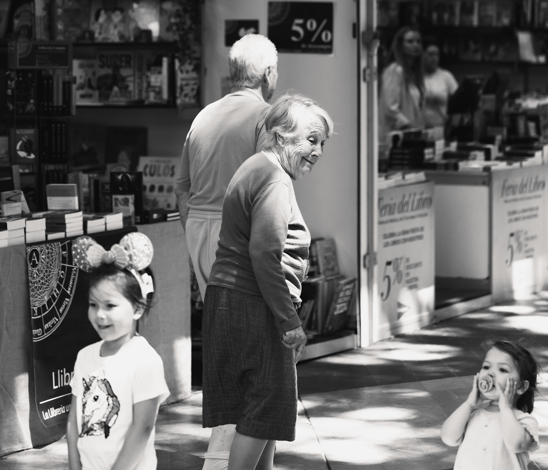 captured on the paseo del borne in palma de mallorca, this black-and-white photograph beautifully juxtaposes generations in a moment of shared space. an elderly woman and man, absorbed in their thoughts, stand near a lively book stall, while young children, brimming with energy and curiosity, move joyfully in the foreground. the contrast between the quiet reflection of the older couple and the vibrant playfulness of the children encapsulates the essence of life's continuum, highlighting the enduring connections between past and present, wisdom and innocence.