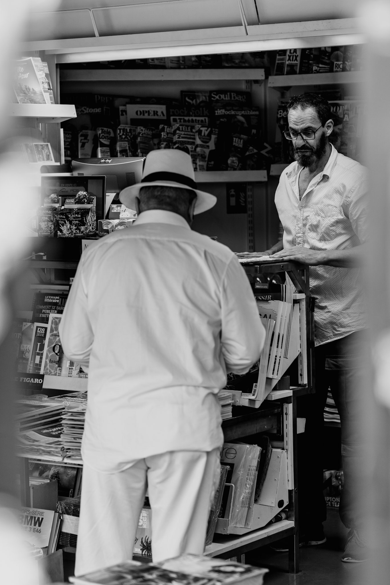 through the frame of modernity, the echoes of history reverberate in the fleeting moment of a newspaper purchase. an everyday ritual captured amidst the storied charm of place des lices in saint-tropez. the juxtaposition of the man's contemporary attire against the backdrop of a traditional kiosk underscores a dialogue between the past and present, bridging generations through printed words.