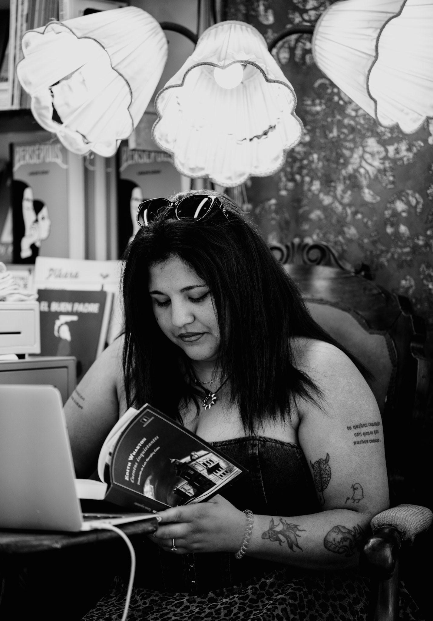 captured at paseo del borne during a book fair, this photo depicts a woman engrossed in a book amidst a display of lamps and books. Booksellers had set up their stalls along the promenade, offering a wide variety of books for sale. The woman, with sunglasses perched on her head and tattoos on her arms, reads intently, immersing herself in the literary world around her. The black and white tones of the image enhance the timeless atmosphere and the quiet moment of contemplation and escape into the world of books.