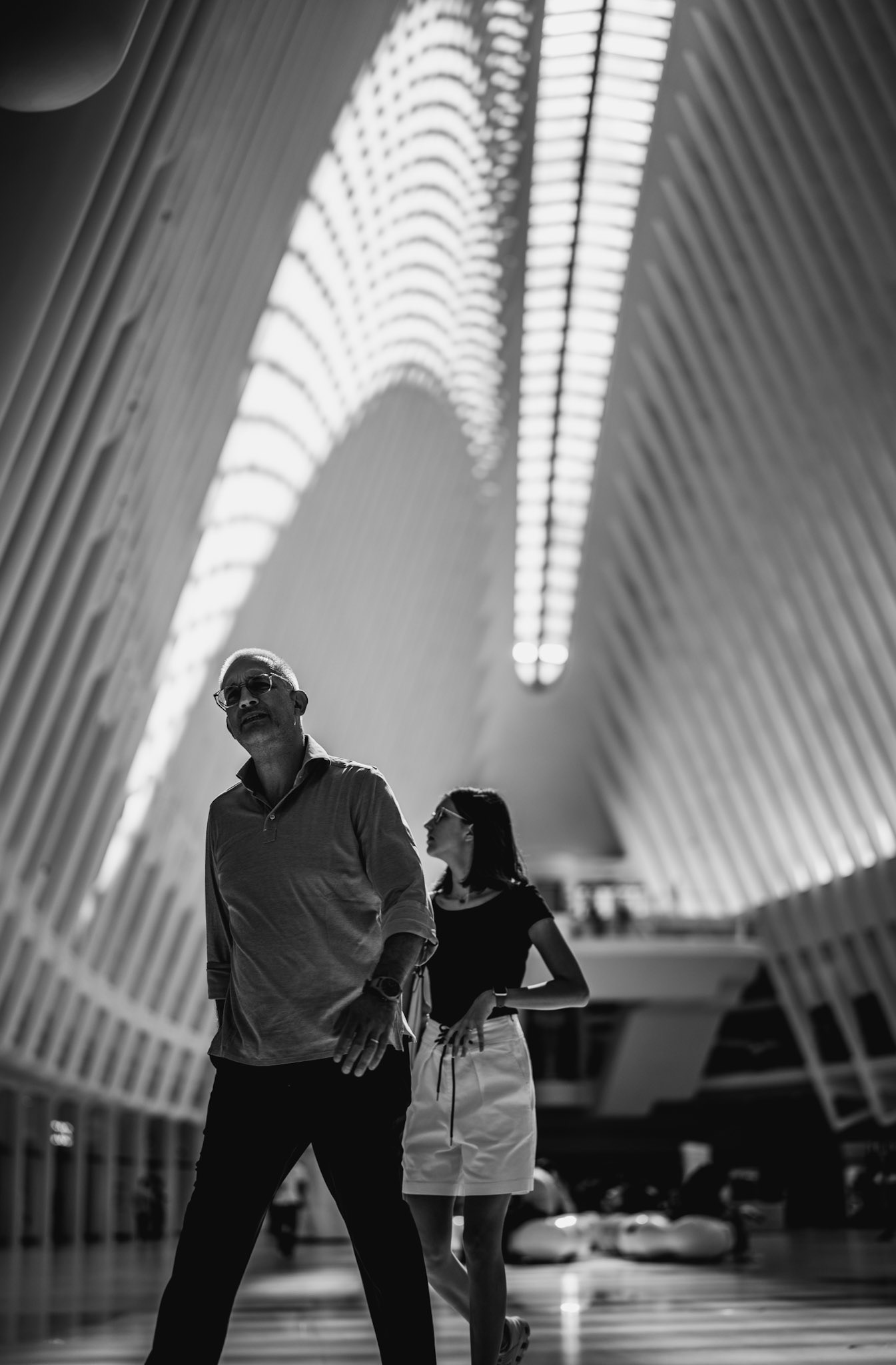 in the heart of new york’s oculus, light weaves through steel like a modern cathedral’s embrace. two figures, grounded yet small, walk in the space, dwarfed by the sweeping curves and sheer height above. they move in unison, caught in the rhythm of this architectural marvel, each step mirroring the structure’s vastness around them.