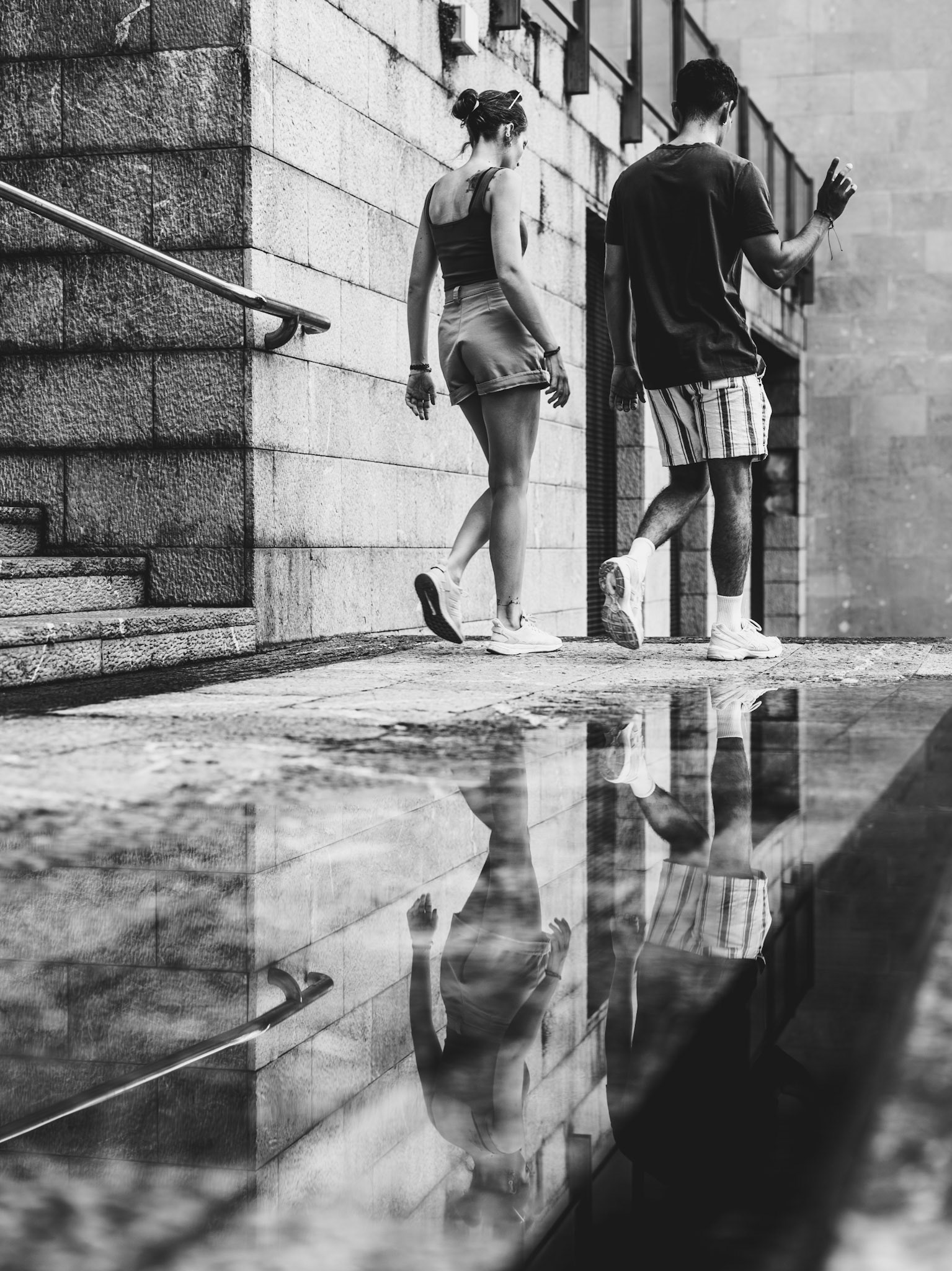two tourists walk down the open stairs that connect the plaça mayor with la rambla, their reflections softened by a shallow pool of water. their footsteps echo gently off the stones, as they leave the busy square above, likely heading to the shade of la rambla’s tree-lined promenade, where rows of poplars offer a cool respite. the steps, weathered and full of history, guide them down into the quieter streets below, perhaps toward a peaceful bench where they can relax and watch the city pass by. it’s a small moment, yet filled with the quiet pace of exploration.