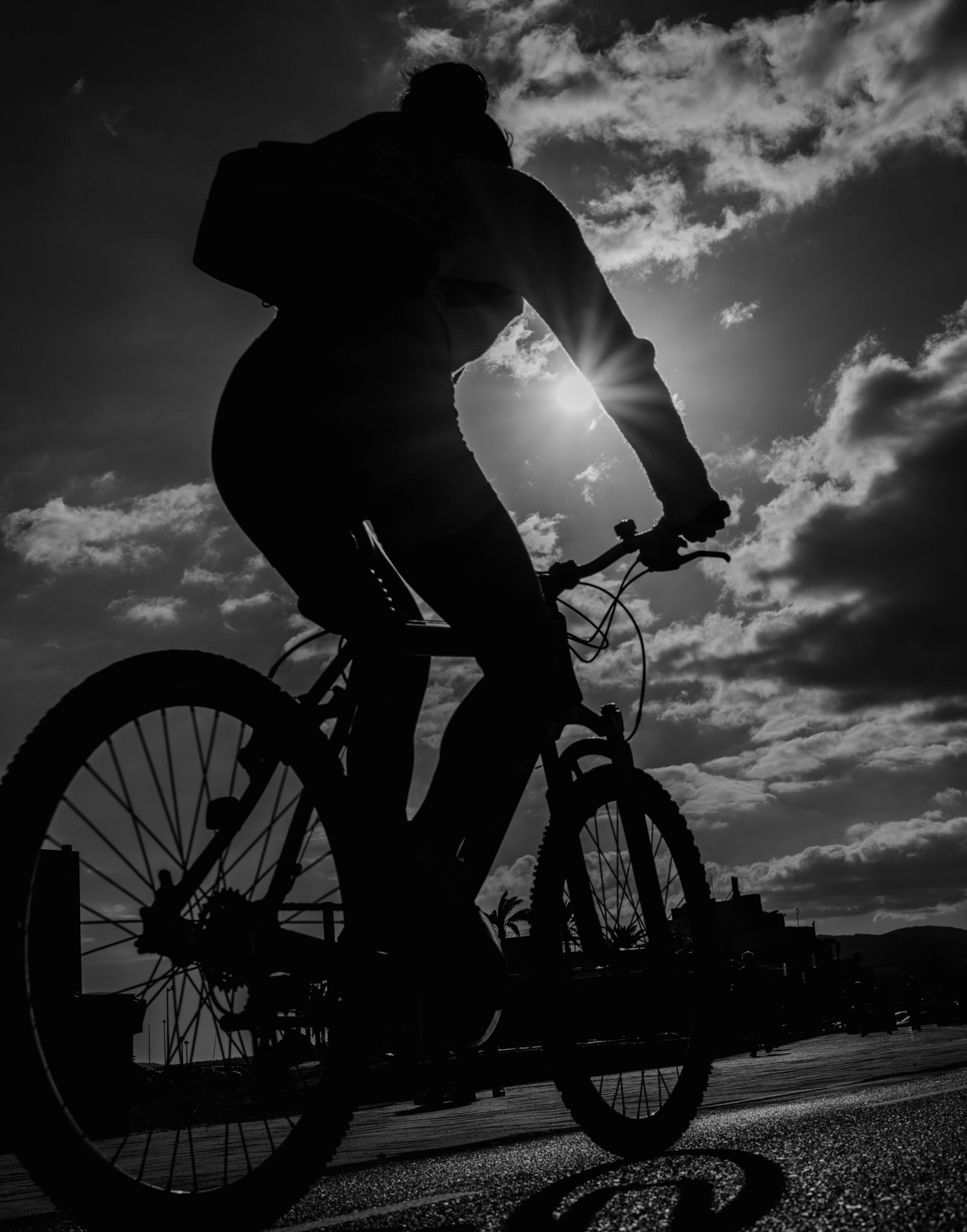 pedaling along the portixol waterfront, the cyclist becomes a bold silhouette against the canvas of a fading day. the sun, a fiery orb, dips low, threading light through the spokes of a turning wheel. clouds scatter across the sky, spectators to this daily race against twilight. the world is reduced to stark contrasts and simple truths in this moment: the eternal cycle of day and night, the solitary figure against the elements, and the pure, unfettered joy of movement. every turn of the wheel is a fleeting dance with the sun's last rays, a pursuit of the horizon as it blushes with the promise of night.