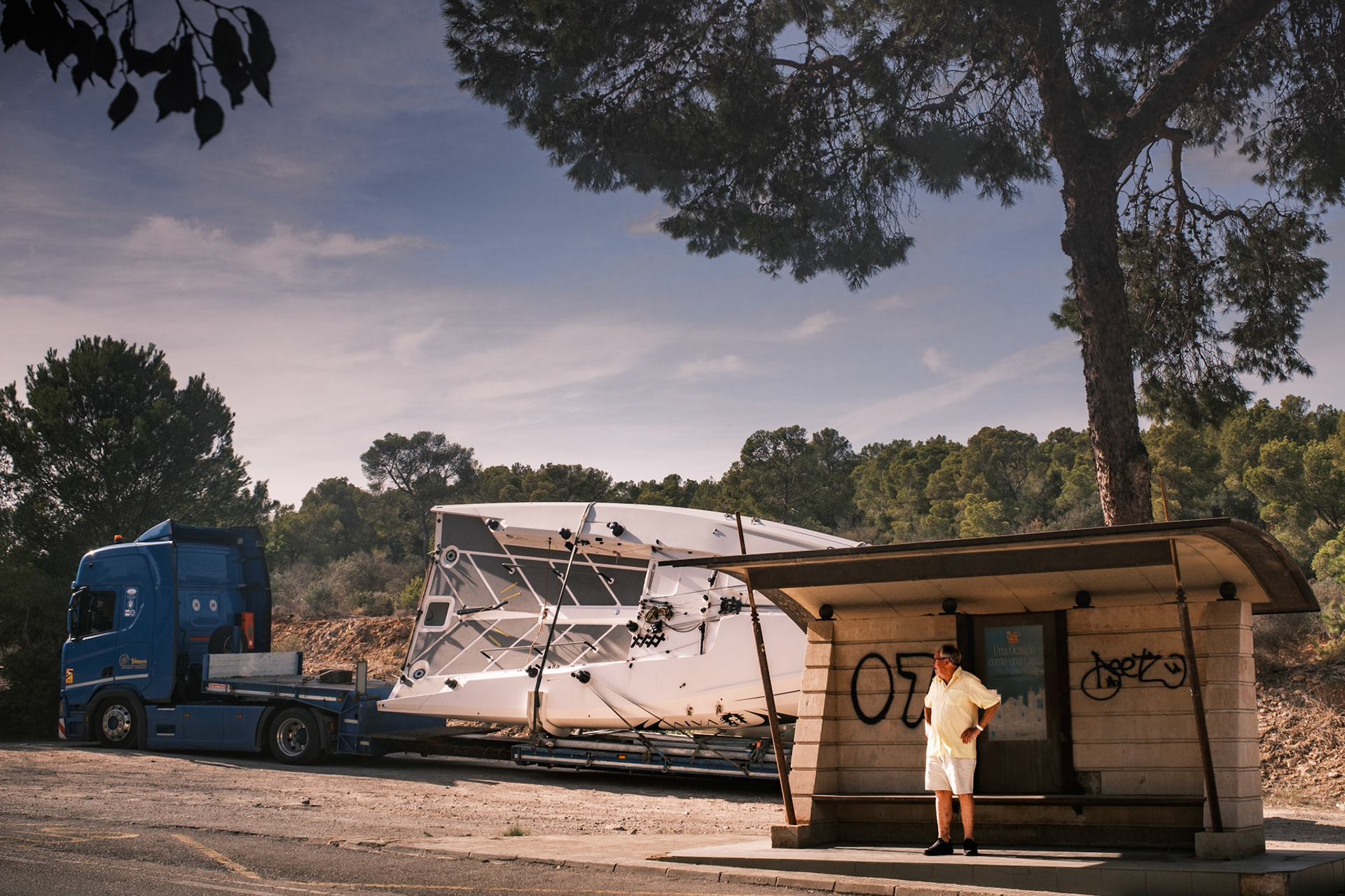 in a scene that feels almost like an illusion, this man waits calmly at what appears to be a bus stop. but is it really a bus stop? and why is there such a massive sailboat parked right beside him, overshadowing everything else in the frame? the boat, the man's casual stance, and the strange pairing of these elements ask a quiet question: is he really waiting for a bus or something else entirely? or perhaps this is just a place where nothing is as it seems, where time stands still, and the bus will never arrive.