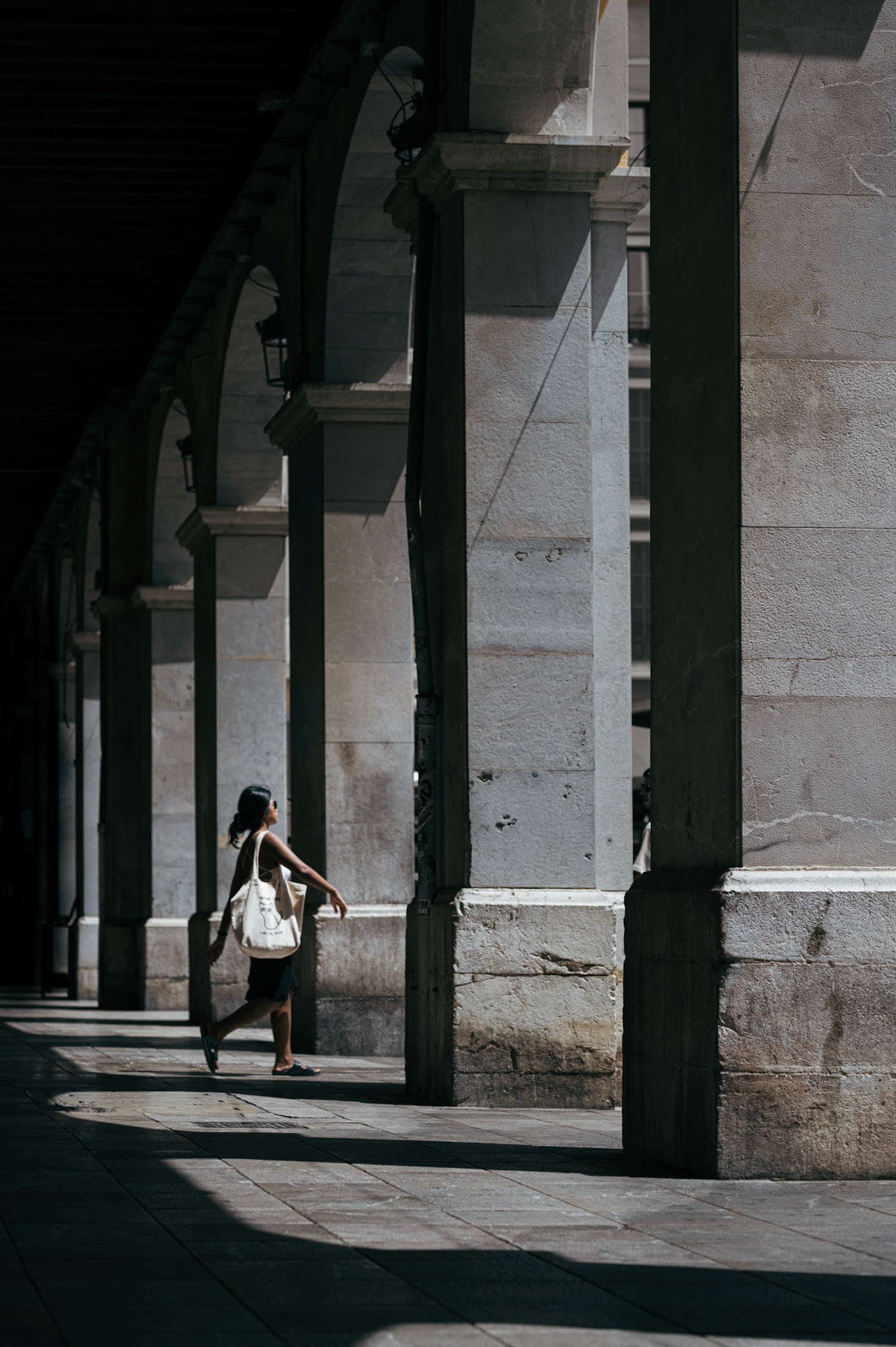 captured at plaça major in palma de mallorca, this image showcases a woman walking through the sun-dappled arches. the play of light and shadow on the stone columns and pavement creates a dramatic backdrop for her purposeful stride. her tote bag and casual attire contrast with the historic architecture, blending modern life with timeless surroundings. this photograph captures a moment of movement and stillness, emphasizing the interplay of light and shadow in a vibrant urban setting.
