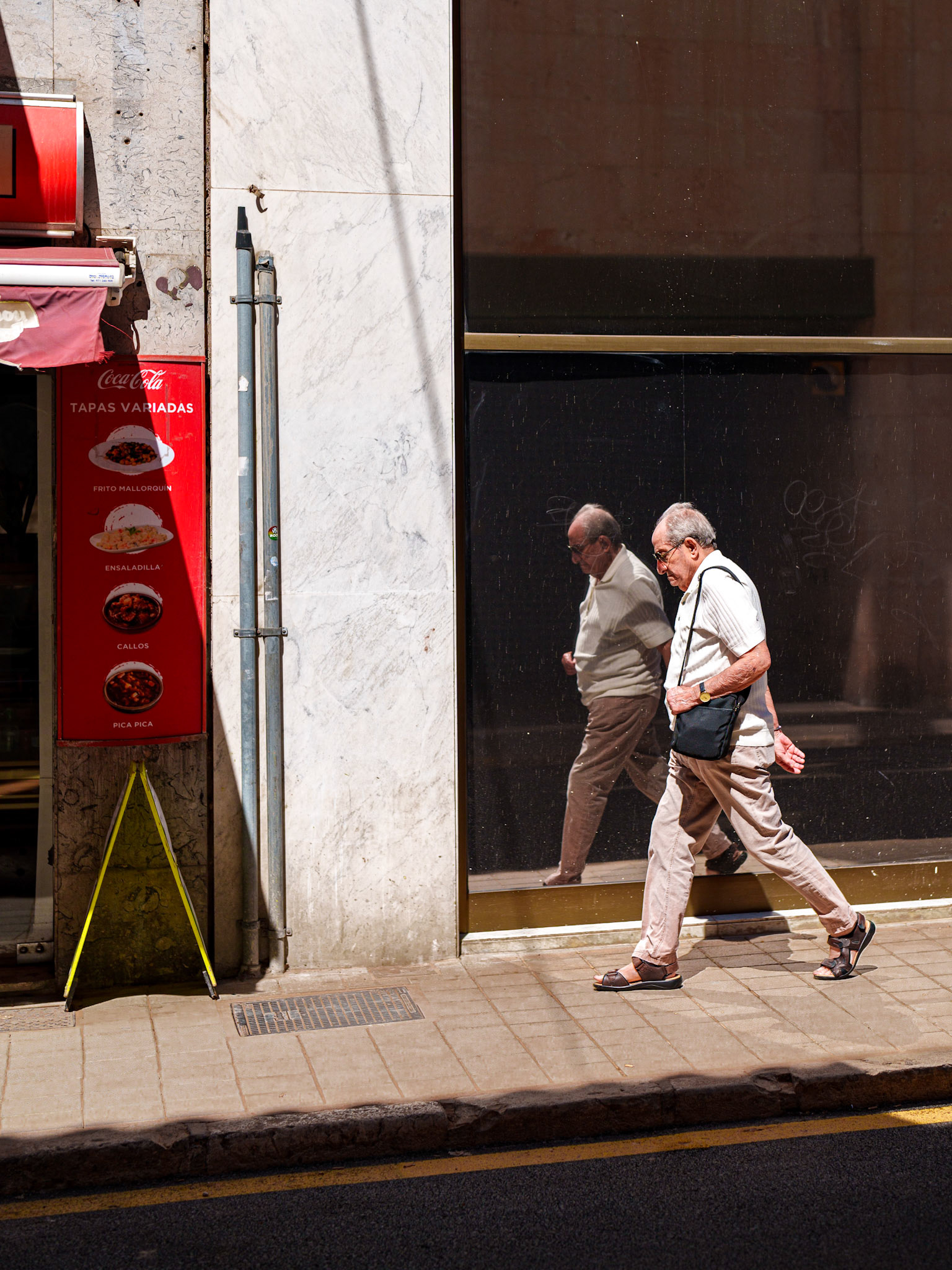 an elderly man walks past a dark, reflective window on a sunny street in palma de mallorca. his reflection mirrors his stride, creating a visually compelling symmetry. the play of light and shadow enhances the composition, highlighting the textures of the stone facade and the man's contemplative expression. this image captures a moment of solitude and reflection in the midst of urban life, where the mundane becomes poignant.