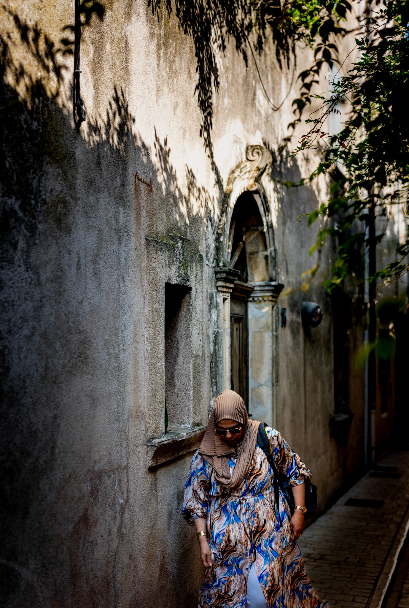 in the heart of saint-tropez, the early morning sun casts intricate shadows upon timeworn walls. a woman, draped in a colorful, flowing dress, moves through the narrow alley of rue etienne berny, her head bowed in contemplation. the play of light and shadow creates a delicate lacework on the rustic facade, narrating stories of the past. every step she takes seems to echo with whispers of history, while her vibrant attire adds a touch of modernity to the ancient street. this juxtaposition captures the essence of a place where time stands still, yet moves forward with every passing day.