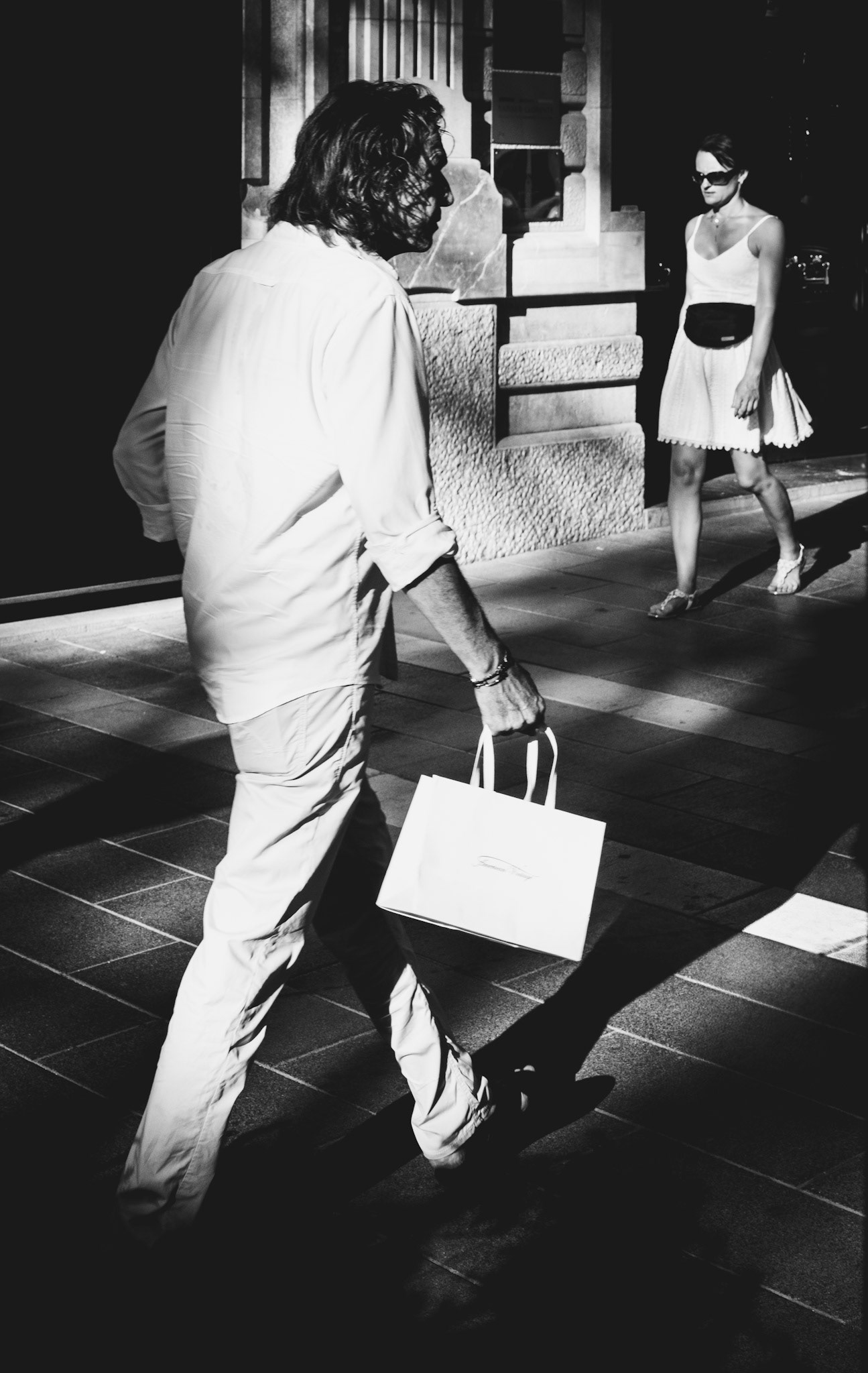 captured on a warm evening along palma’s elegant paseo del borne, this photo juxtaposes two passersby dressed in all-white attire. the contrast is accentuated by the play of light and shadow, with the man’s white shopping bag further emphasizing the monochromatic theme. the deliberate composition, with its sharp contrasts and the parallel paths of the subjects, captures a fleeting moment of urban life, embodying both simplicity and sophistication in a single frame.