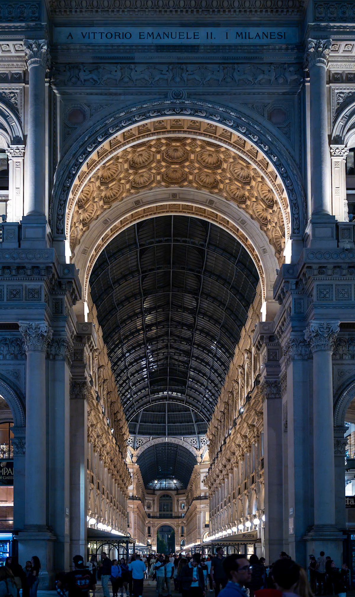 this photograph showcases the grand portal of the Galleria Vittorio Emanuele II, opening onto the vibrant Piazza del Duomo in Milan. The striking contrast of light and shadow accentuates the intricate reliefs and opulent architecture, a testament to the grandiosity of Italian design. Under this ornate canopy, visitors and locals alike traverse, small figures against the monumental backdrop, partaking in the age-old tradition of the evening passeggiata. It’s a place where history is etched into every detail, and modern life flows beneath the watchful eyes of a bygone era.