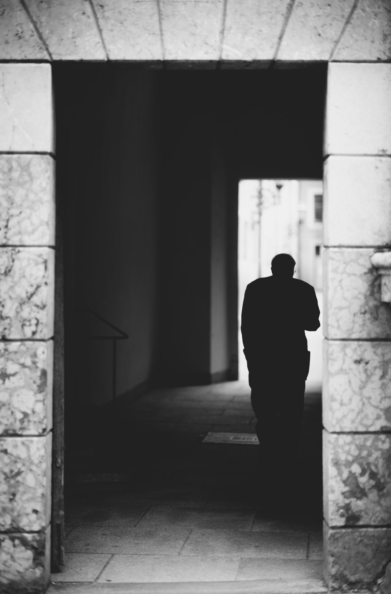 captured in munich at the hofgarten, this image shows a solitary figure walking through a shadowed archway, emerging into the light beyond. the black and white composition emphasizes the contrast between darkness and light, creating a powerful sense of transition. the worn stone of the archway frames the scene, adding a timeless quality. this photograph evokes a sense of mystery and contemplation, as the figure moves from the shadows into the light, symbolizing a journey through time.