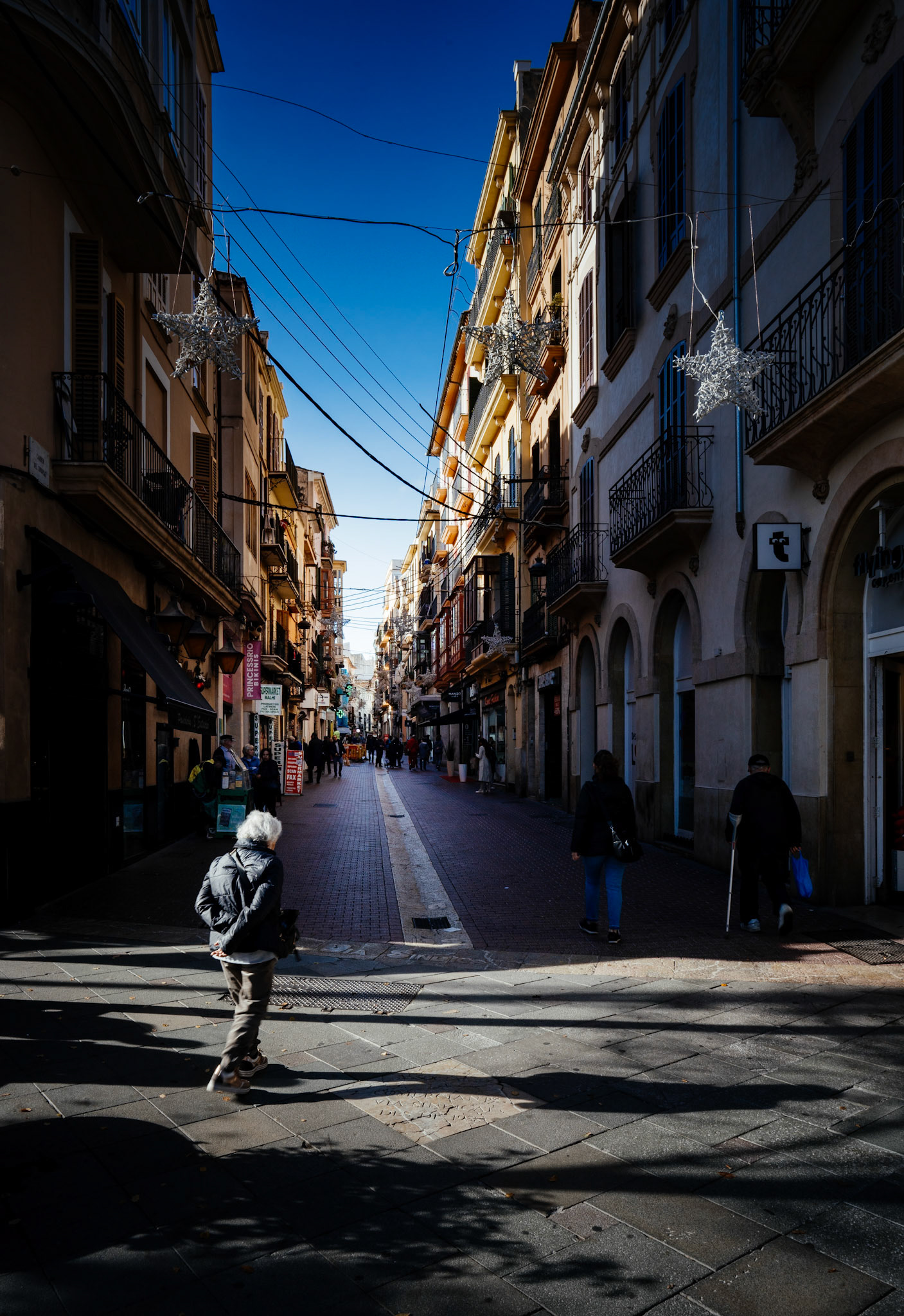 in the heart of palma, the calle sindicat is bathed in the dramatic light of a sun that plays hide and seek with the city's architecture. a solitary figure traverses the street, their shadow stretching long and mingling with the patterns of the pavement. above, festive stars hang, remnants of celebration, contrasting with the everyday scene below. it's a moment suspended between the lingering spirit of festivity and the steady rhythm of local life.