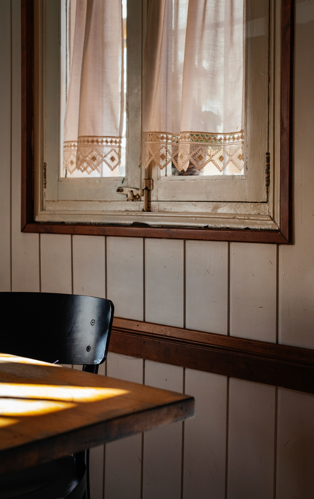 in "el viejo y el mar," sunlight bathes a solitary chair beside a rustic table. the window, adorned with lace curtains, frames the noon light, creating a serene tableau within the walls of palma de mallorca's vibrant district of pere garau. an air of calm anticipation fills the cozy space.