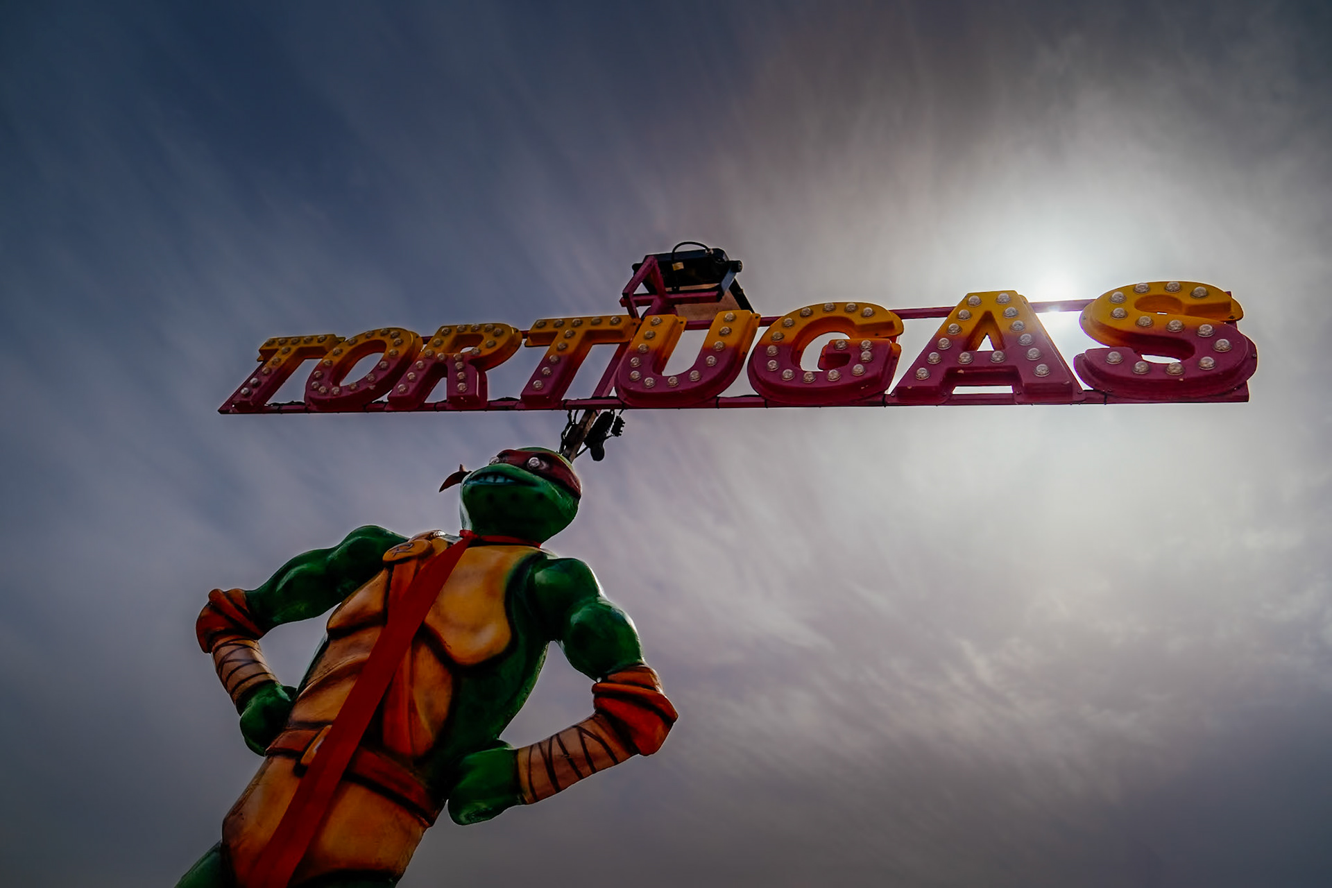 against the backdrop of a late afternoon sky, the sun plays hide and seek behind an iconic figure reimagined as a sentinel of childhood joys. the 'tortugas' sign, a beacon of festivity, crowns the scene in bold, carnival colors, echoing with the laughter of the fiera del ram. there's a sense of movement in the stillness, a dance of light and shadow, as if the figure is about to leap into the throng of carousel music and spinning lights. it's a moment suspended, a clash of fantasy and reality, where stories from our youth stand tall against the infinite canvas of the sky.