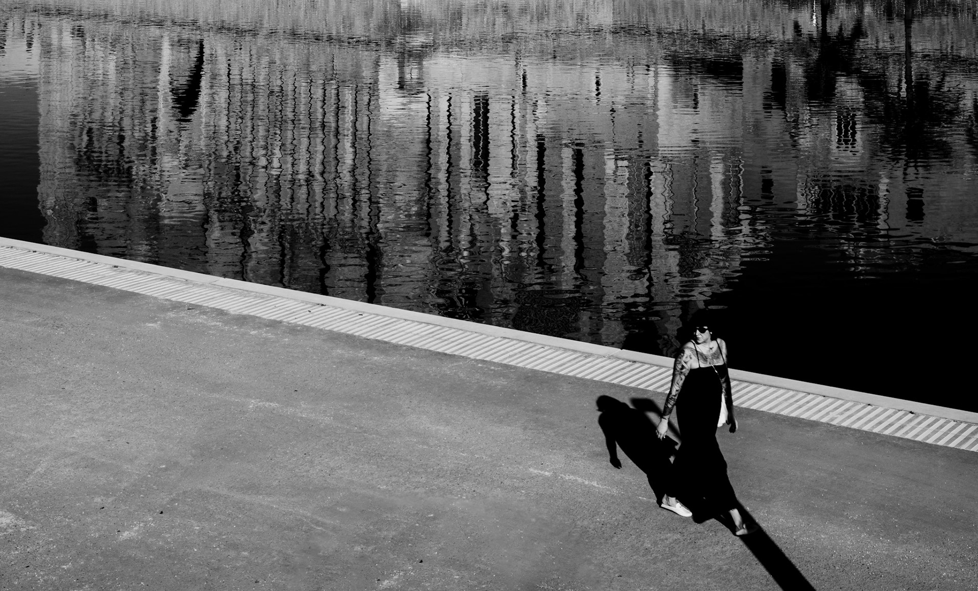 In the serene setting of Parque del Mar in Palma de Mallorca, the still waters create a perfect mirror for the majestic Cathedral of Palma. A solitary figure walks by, her silhouette casting a sharp contrast against the shimmering reflections. This fleeting moment captures the tranquil essence of the city's waterside.