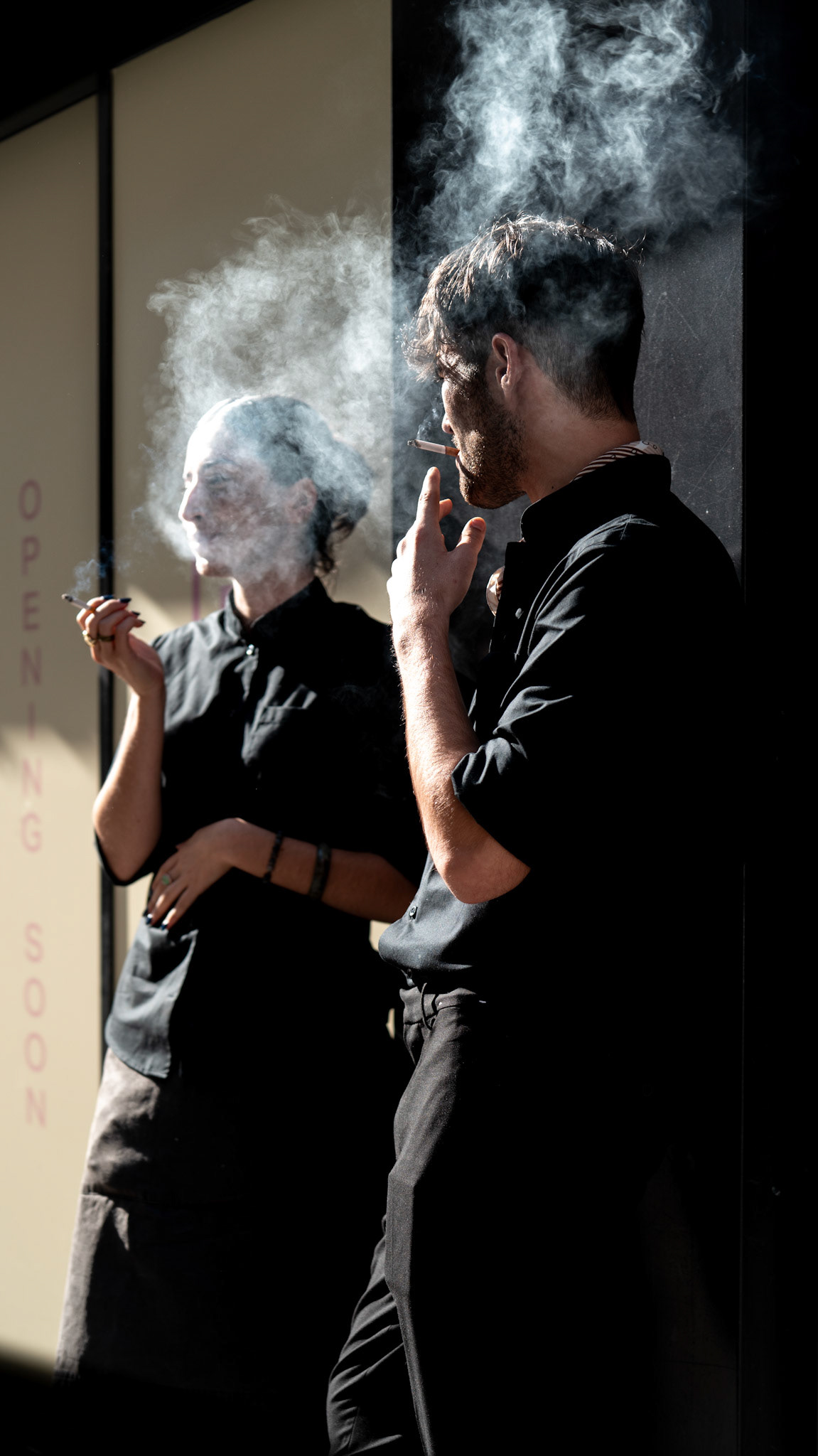 in the heart of paris, on a quiet corner of rue du bac, two workers take a brief respite from their daily routine. framed in the golden light of the afternoon, smoke swirls around them, creating a dreamy haze that contrasts with the crispness of their black uniforms. the scene feels almost timeless—a shared moment of calm amidst the bustling city streets. the way the light and shadow play on their faces adds depth to the image, bringing out the contemplative expressions of each figure, making you wonder what thoughts linger behind the smoke.