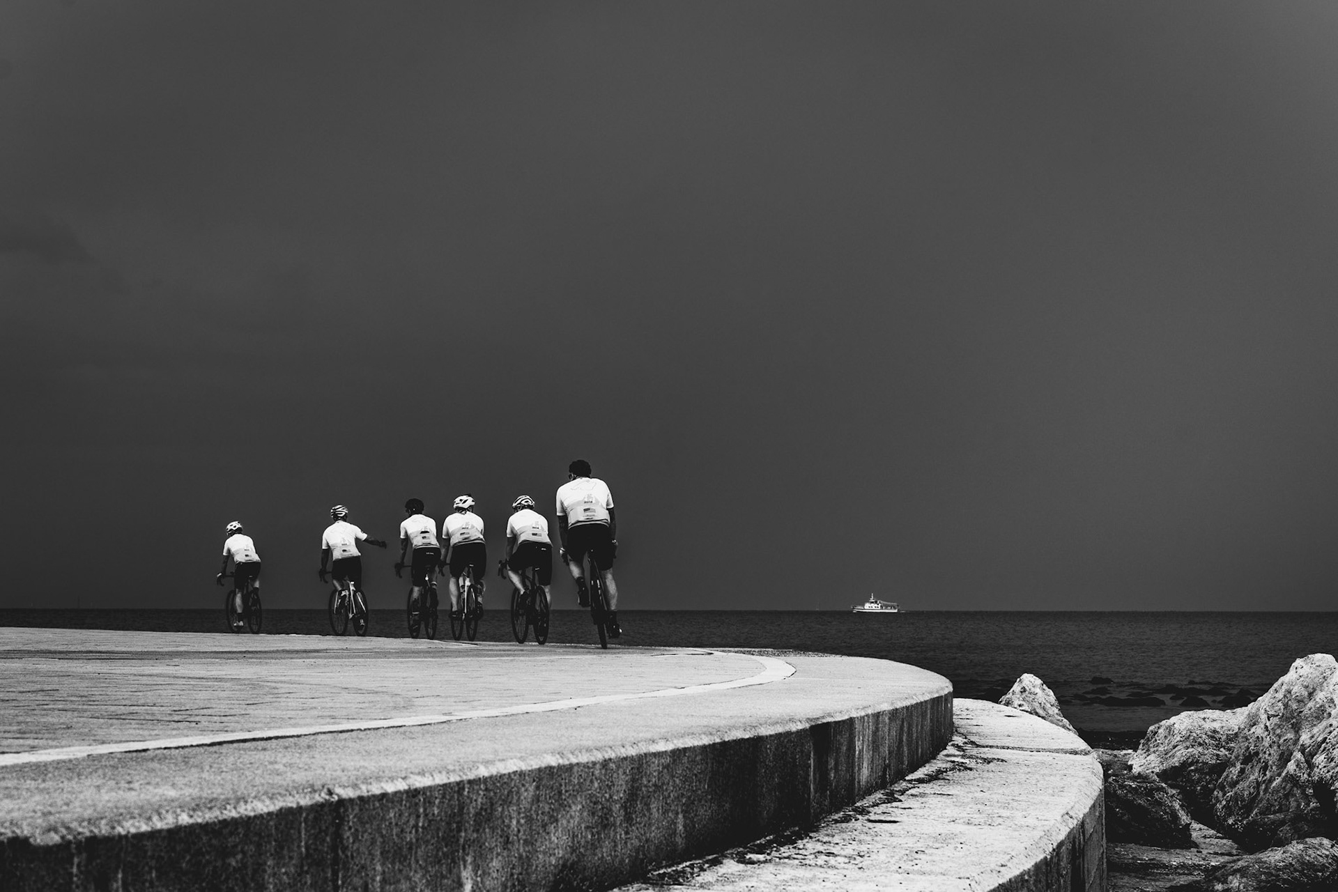 under a gray, heavy sky, a group of cyclists follows the curve of the pier, riding towards the distant horizon where a lone boat drifts on the sea. the strong lines of the pier lead the eye into the open ocean, connecting the movement of the riders with the stillness of the water. a striking juxtaposition where motion meets calm, and the vastness of the sea contrasts with the human determination to explore its boundaries. the scene suggests a journey with a destination still uncertain, yet undeniably present.