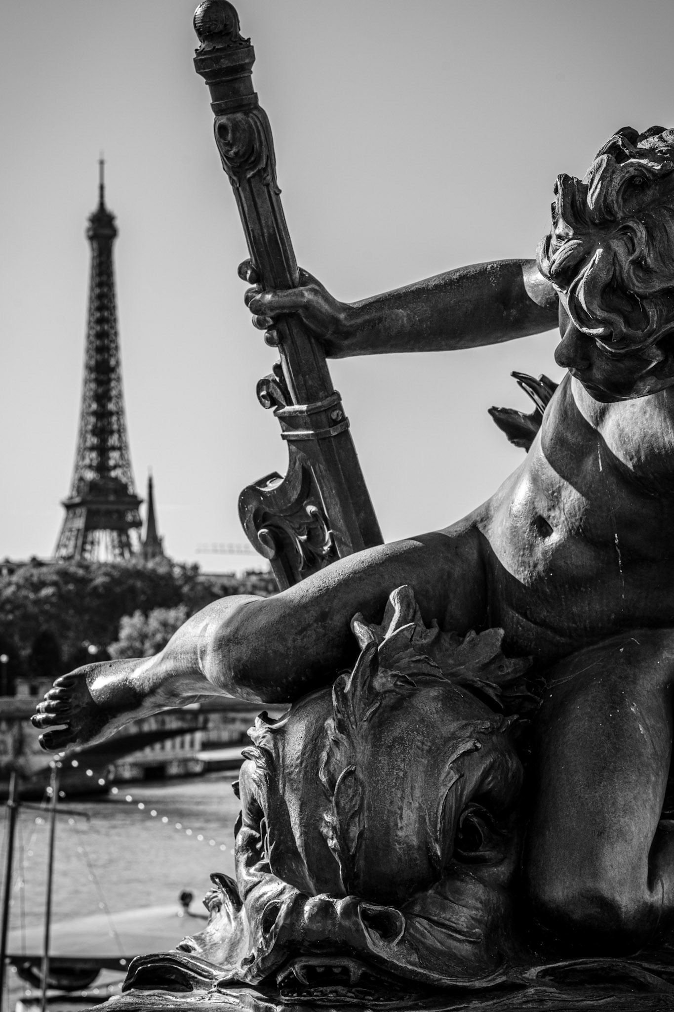 this image captures the details of one of the ornate cherubs on the pont alexandre iii bridge in paris, a masterpiece of beaux-arts architecture spanning the seine. commissioned for the 1900 world’s fair, this bridge symbolizes french-russian friendship and is widely considered one of the most beautiful bridges in the world. each cherub and nymph adorning it tells its own story, embodying the themes of art, music, and maritime power.the cherub here seems to represent the spirit of music, clutching what looks like an ornate lyre. positioned against the iconic eiffel tower, the sculpture draws a unique connection between paris’s architectural beauty and its artistic soul. the contrast between the worn bronze textures of the statue and the elegant tower in the distance gives a sense of depth and narrative, as if time stands still here, blending the classical and the modern.