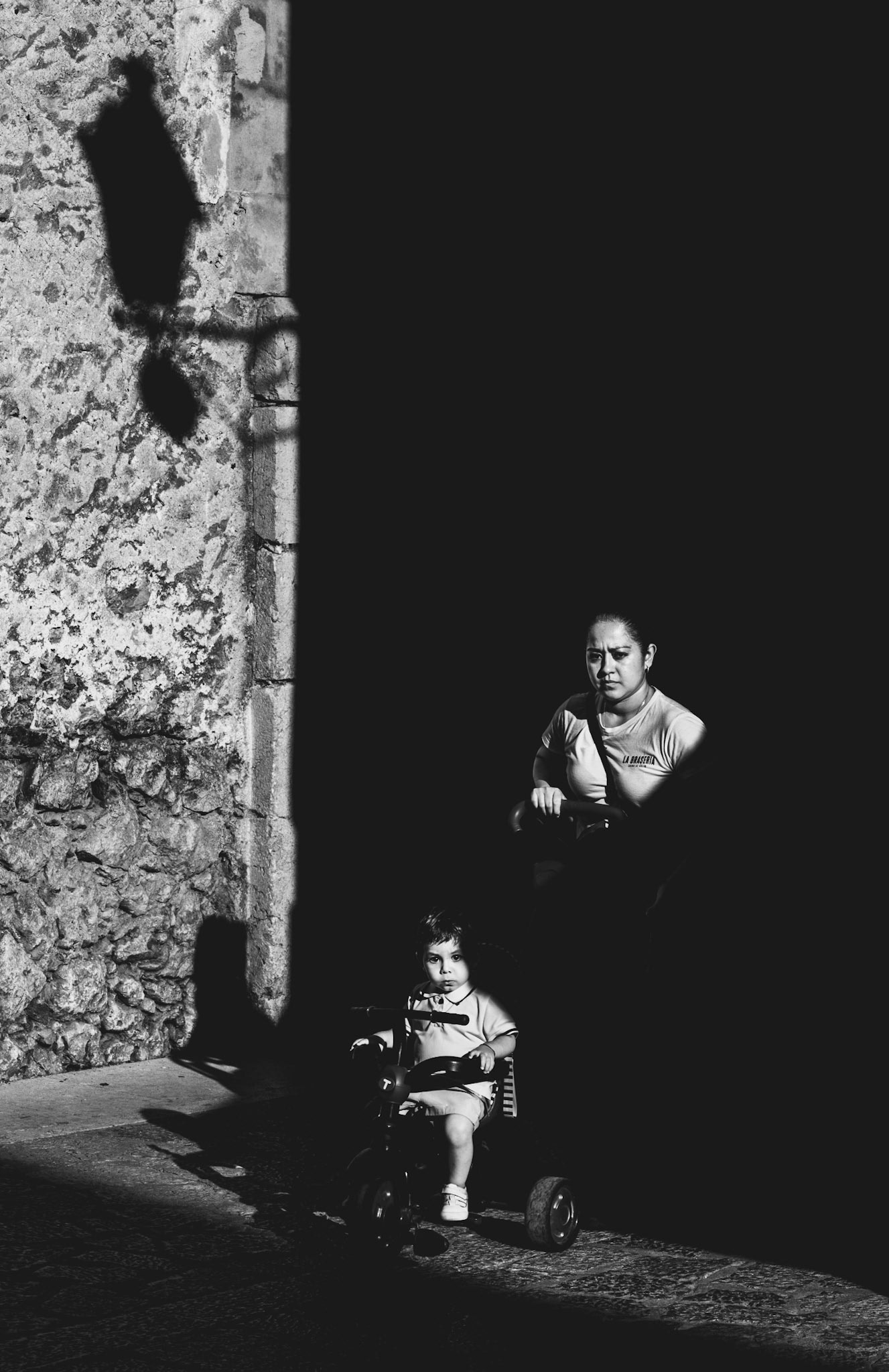 in the narrow streets of pollença’s old town, a harsh beam of sunlight illuminates just a part of the scene, creating a dramatic tension between light and darkness. a woman pushes a child on a tricycle through the alley, both caught in the bright light, while the rest of the world around them dissolves into deep shadows. the child's face shows a mix of curiosity and quiet seriousness, while the woman seems absorbed in her thoughts, framed by the sharp contrast of the light. the silhouettes of the streetlamp and the rough textures of the old stone wall add a sense of mystery, inviting the viewer to imagine the untold stories behind this moment. a photograph that captures the intensity of everyday life in a fleeting second, set against the timeless backdrop of pollença’s historic streets.