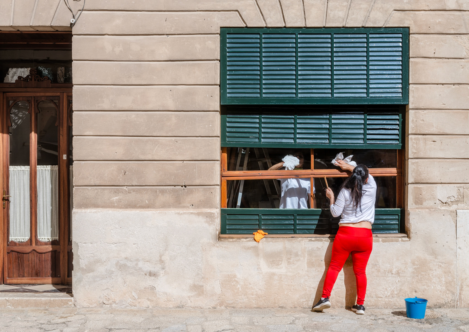 nestled in the gentle light of pollença's old town, an everyday scene unfolds before an age-old window. two women, clad in white and red, stand out like vibrant strokes of paint yet blend into the historical tapestry. they work in unison, the window their shared charge, tending to the legacy before them. half-open, half-closed, it captures their exchange—not of words, but of movements, a shared rhythm in the act of cleaning and renewal. a simple bucket stands witness to the unassuming nature of this scene, rich in history and community.
