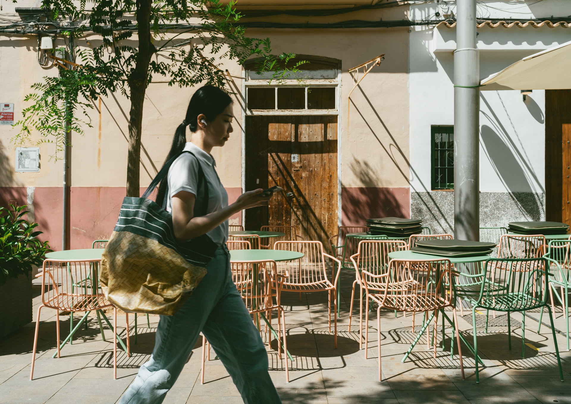 in the warm embrace of the afternoon sun, a solitary figure walks past a quaint cafe in palma de mallorca, her focus absorbed by the screen in her hand. the pastel-colored chairs cast intricate shadows on the pavement, creating a dance of light and form. the rustic charm of the weathered walls behind her tell stories of time and history, as the modern world moves forward in stride. this moment, a blend of past and present, captures the essence of life's fleeting yet beautiful intersections.