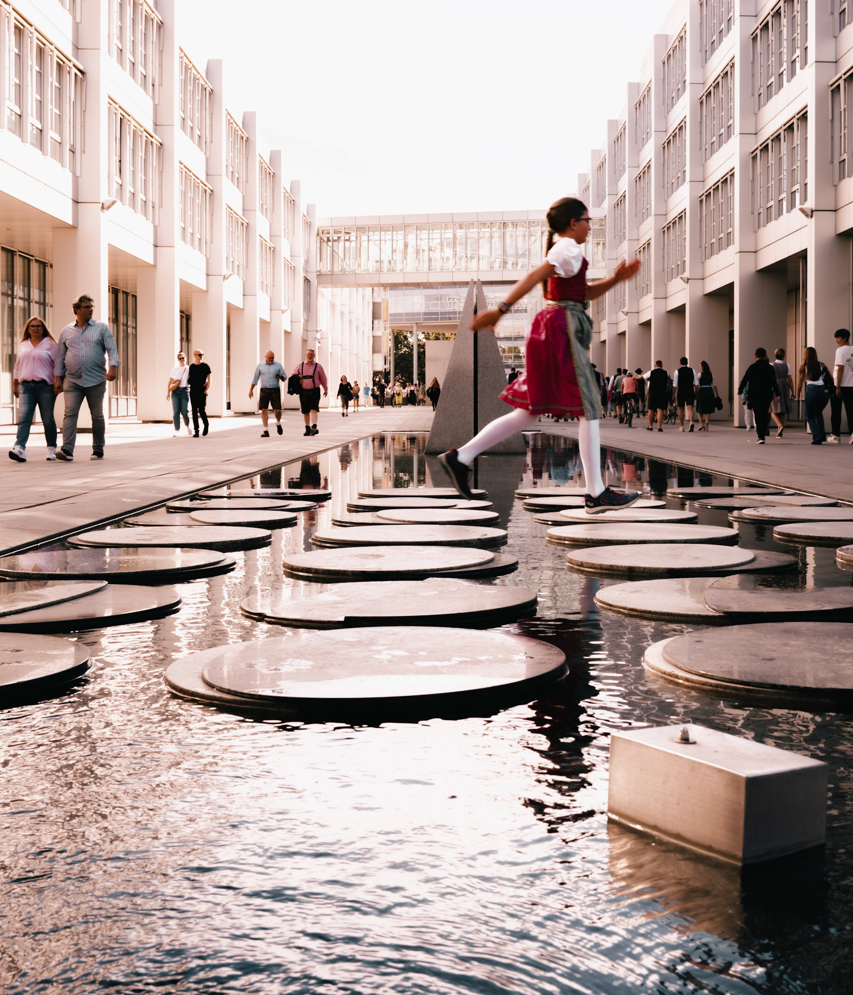in munich, amidst the bustling energy of oktoberfest, a little girl in a vibrant "tracht" is caught mid-motion, leaping over water features. her lightness of being contrasts with the urban landscape of the city. the scene is a delightful blend of heritage and modernity, a dance of tradition in contemporary times. as the city indulges in its annual festivities, this fleeting moment captures the spirit of oktoberfest – joyous, vibrant, and steeped in bavarian culture. her movements seem to echo the heartbeats of munich, pulsing with life and tradition.