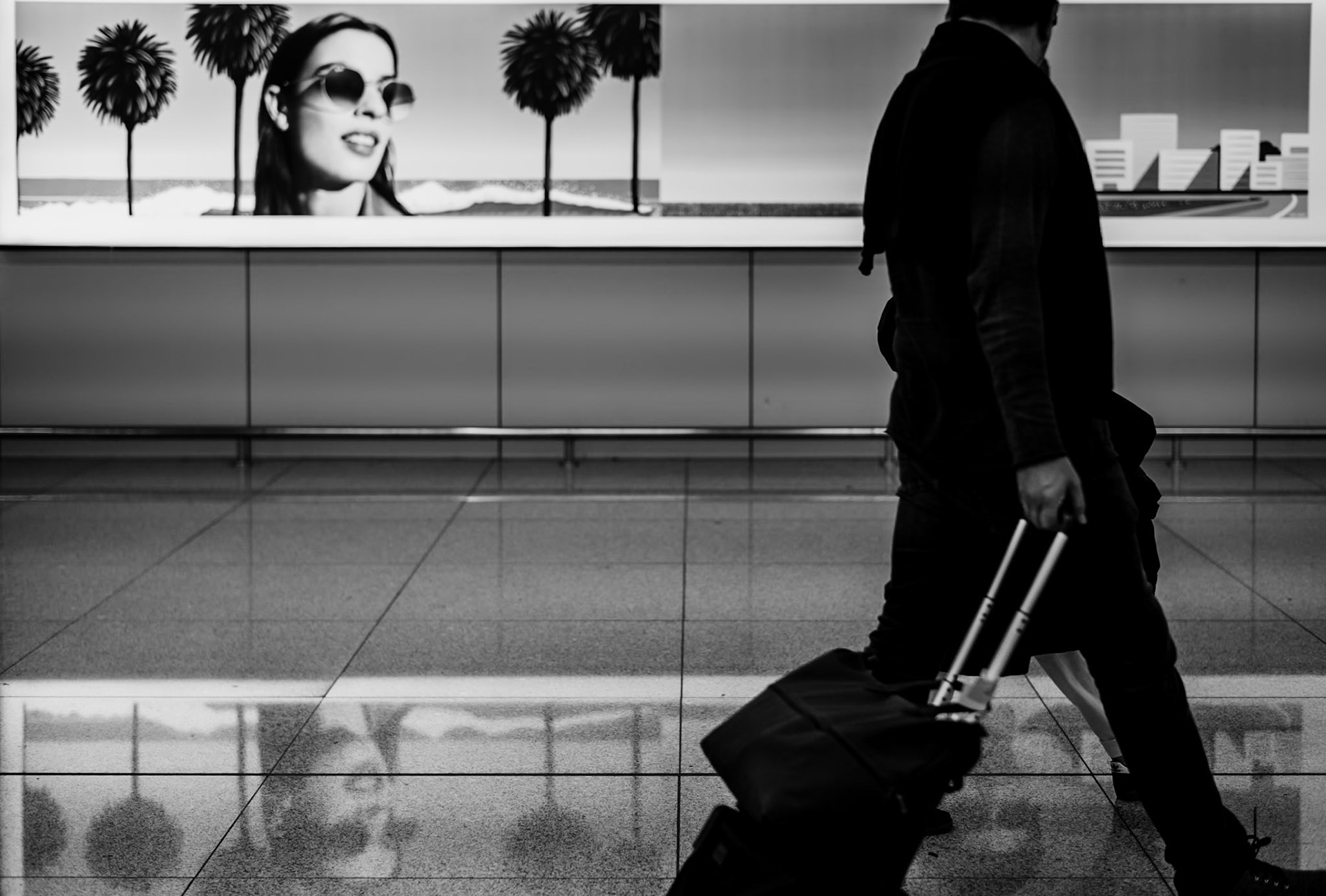 In the transient stillness of Palma's airport, a juxtaposition emerges. The laughter of the sun-kissed face, immortalized in a billboard, contrasts with the hurried stride of a traveler. Here, amidst the flux of departures and arrivals, summer whispers its tales through the still imagery amidst the ceaseless motion of passersby. This scene, captured in monochrome, feels like a paused narrative — a moment where the warmth of distant beaches echoes against the cool, hard lines of the present. It’s a dance of light and shadow, presence and absence, a fleeting connection between the joy of sunny escapades and the solitude of travel.