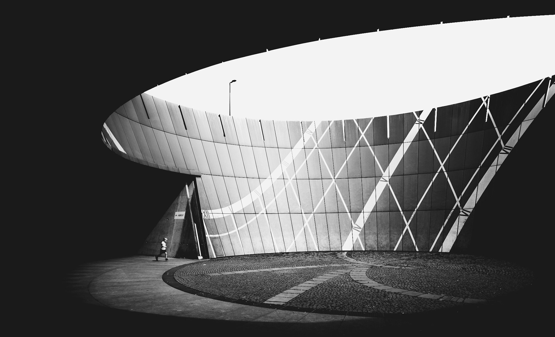 in a moment of stillness amidst an architectural marvel, a solitary figure walks under the vast, elliptical opening of the structure, dwarfed by its scale. the interplay of shadows and reflections creates a dramatic pattern across the curved facade, where lines intersect in a dance of light and geometry. the minimalistic composition and high contrast in black and white elevate the scene to a visual symphony, capturing a fleeting moment in a space that feels both immense and intimate. it's a study of scale, solitude, and the silent dialogue between human and architecture.