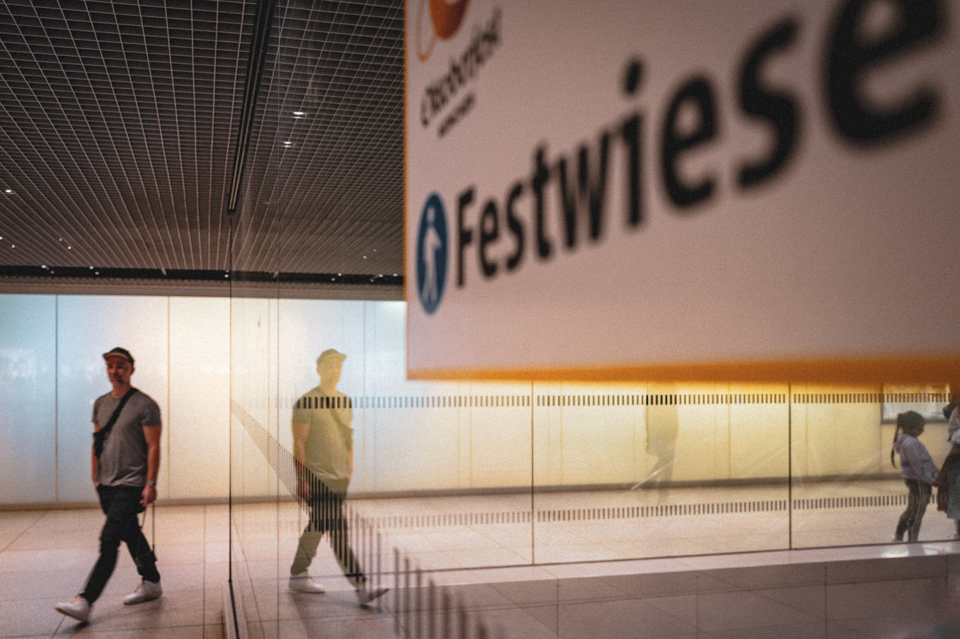 beneath the humming lifeblood of munich central station, a tale of two desires unfolds. a lonesome traveler strides, his path divergent from the beckoning "festwiese" sign, where echoes of oktoberfest revelry call. it's a silent narrative of choices and reflections—literally. his mirrored self, a ghostly silhouette, appears to glance towards the festivities, a doppelgänger drawn to what might have been. in this transient corridor of life's crossroads, every footfall is a story, a singular journey against the tide, a personal odyssey within the city's heartbeat.