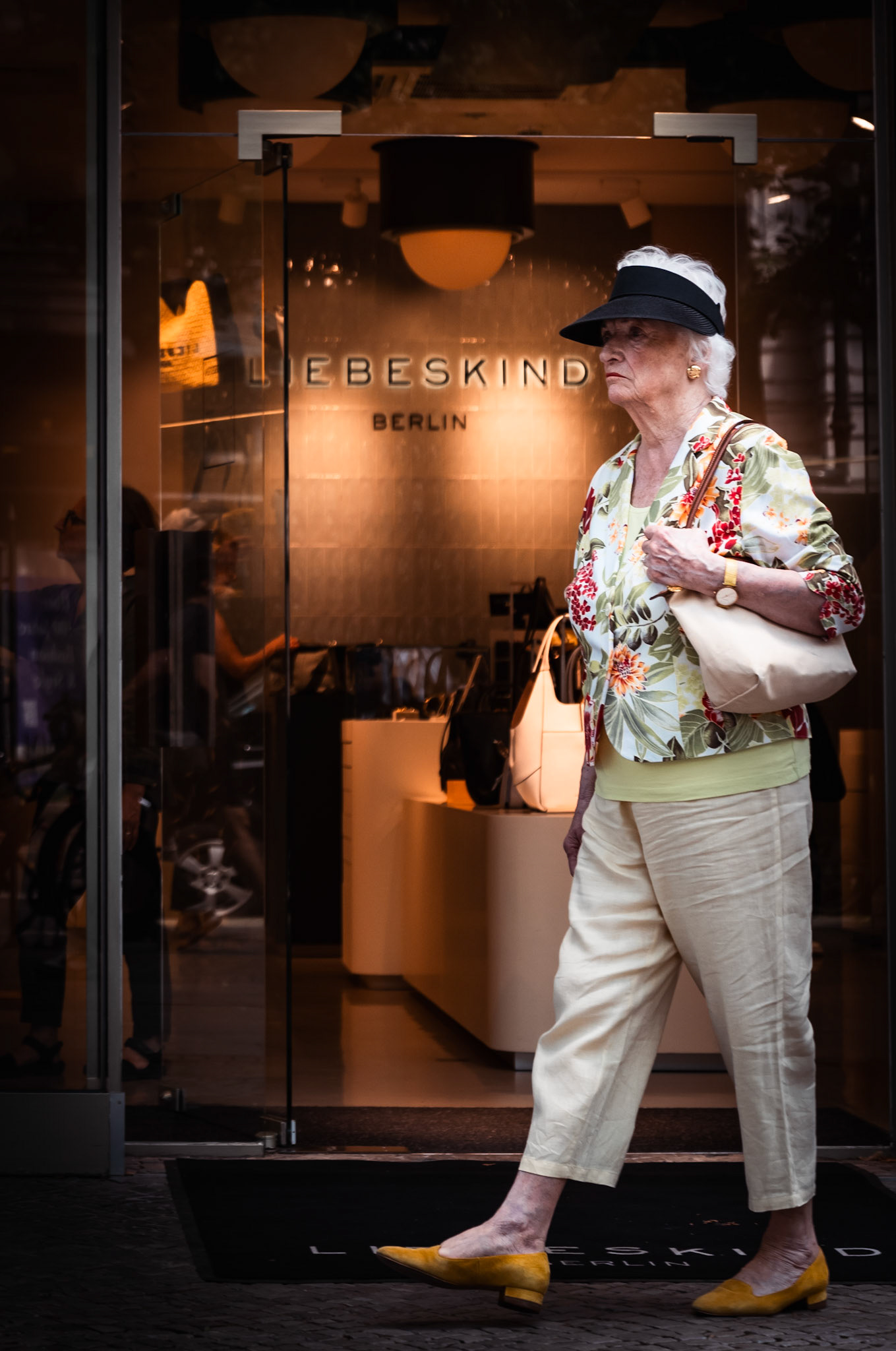 captured on kurfürstendamm in berlin, this photo features an elegant elderly woman examining the storefront of the luxury brand "liebeskind." her sophisticated attire, complete with a stylish hat and floral jacket, complements the upscale ambiance of the setting. the juxtaposition of her timeless grace against the modern retail environment creates a striking visual narrative, highlighting the intersection of tradition and contemporary fashion.