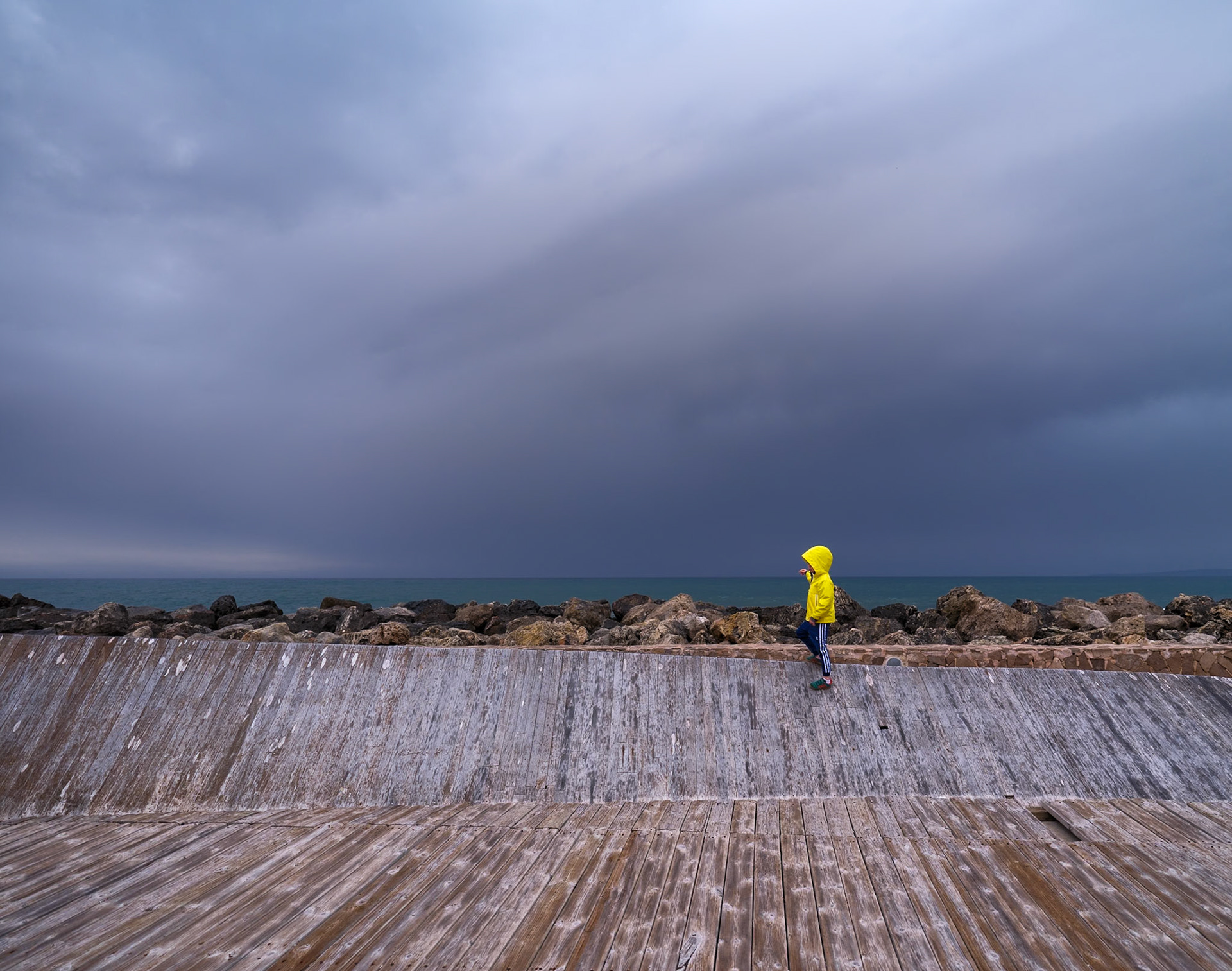 even as evening descends on the harbor promenade of coll d'en rabassa, a child in a yellow slicker stands out against the looming grey of a stormy sky. the wood underfoot bears the weathered stories of countless storms before, a silent testament to resilience. this lone bright figure, small yet defiant, looks out to where the sky meets the sea, a vast canvas of moody blues. it's a scene of contrast and simplicity, where color pops vividly against the muted tones of an impending downpour.