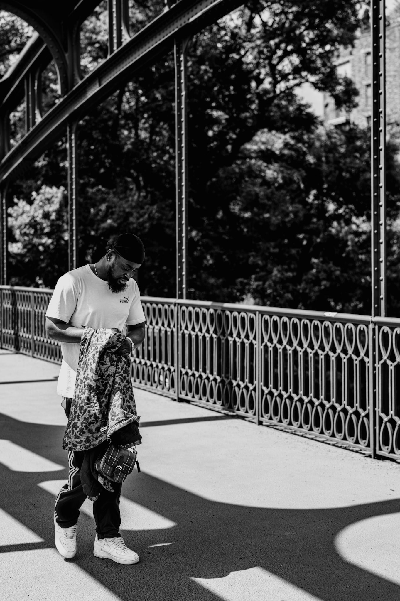 as i wandered through the iron arches of this old bridge, i captured a fleeting moment of solitude. the sunlight poured through the gaps, casting intricate shadows that danced on the pavement. the man, lost in his thoughts, cradled his jacket as if it were a shield against the world. the urban backdrop with its timeless architecture and lush greenery framed his quiet journey, creating a harmonious blend of nature and industry. this image, bathed in monochrome, speaks to the intersection of daily life and the silent whispers of the city’s history.