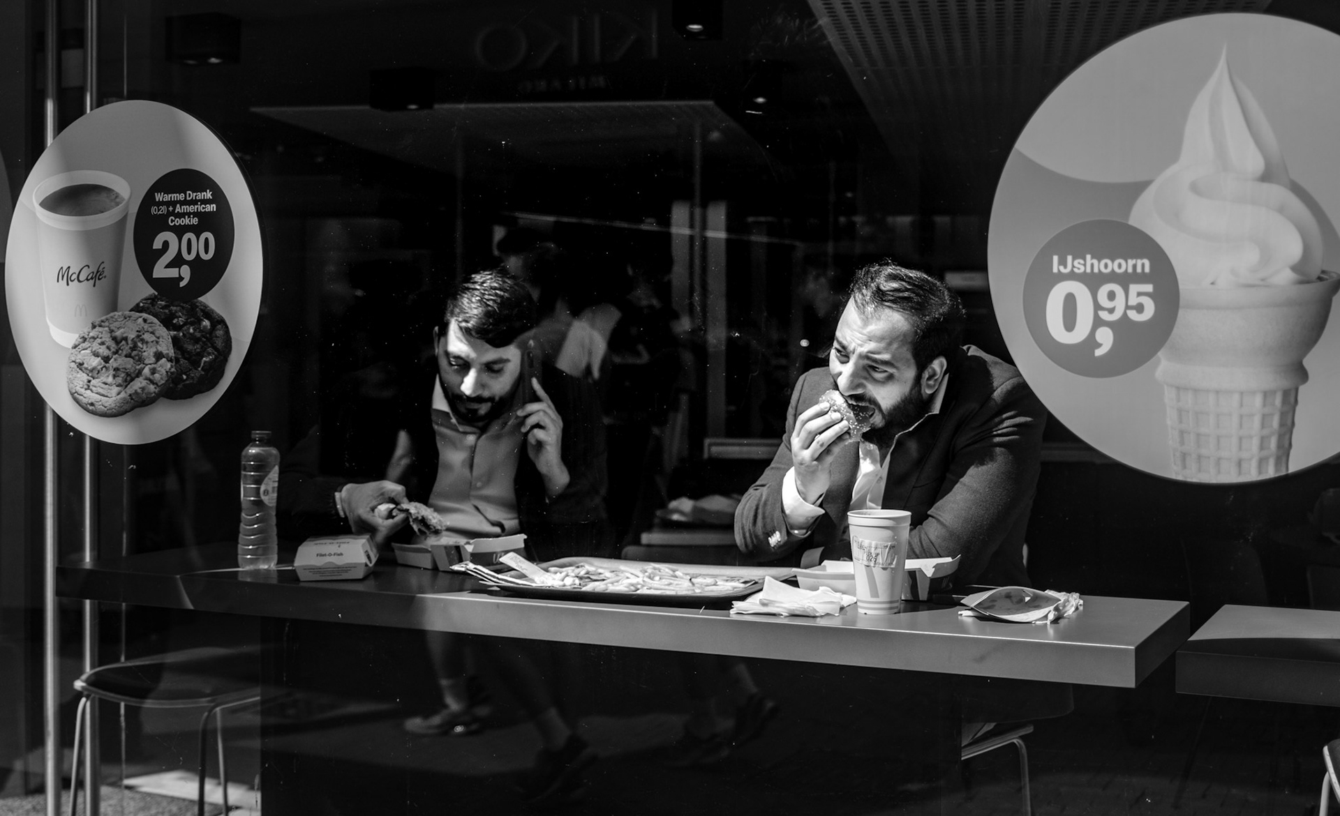captured in the bustling pedestrian zone of amsterdam, this photograph showcases two men enjoying burgers at a well-known fast food chain. one man, deeply engrossed in his meal, bites into his burger with evident satisfaction, while the other, slightly distracted by a phone call, manages a bite amidst the conversation. the reflective glass of the window adds depth to the scene, mirroring the vibrant street life outside and blending the interior moment with the city's lively backdrop. this image beautifully captures the juxtaposition of personal moments and public spaces, highlighting the intersection of modern life and urban culture.
