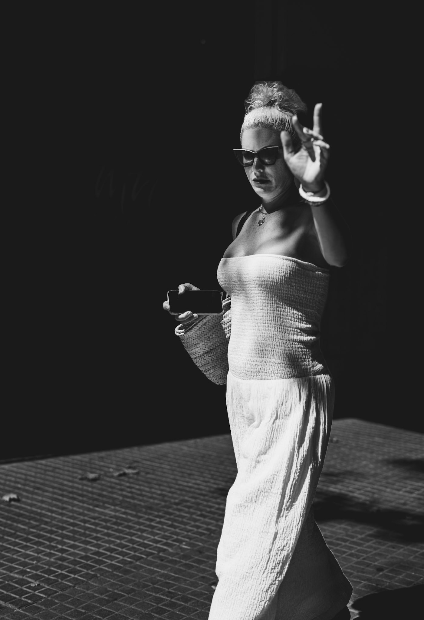 on the sunlit streets of palma de mallorca, a woman in white raises her hand in a gesture of dismissal, her face set in a mask of determination behind dark sunglasses. the stark contrast between the bright light and deep shadows captures the intensity of the moment, emphasizing the strong emotions at play. her body language speaks volumes, a candid snapshot of modern urban life where personal boundaries are asserted amidst the daily hustle.