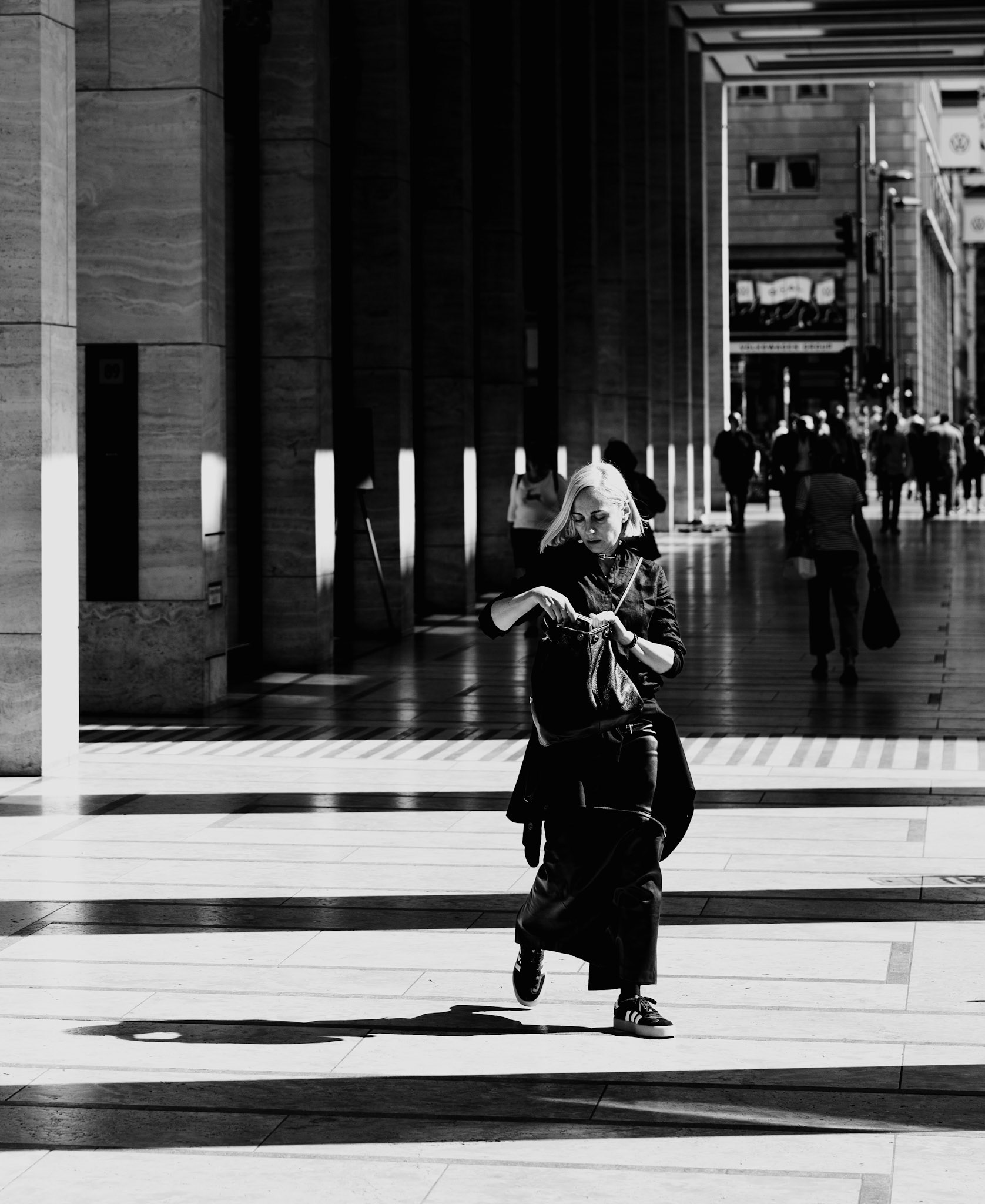 captured on friedrichstraße in berlin, in front of the dussmann das kulturkaufhaus, this photograph plays with the contrast of shadows and light. the woman in the foreground, absorbed in her own world, adds a dynamic element against the backdrop of towering columns and elongated shadows. the black and white composition enhances the urban elegance, drawing the viewer into the rhythm of city life where every shadow tells a story.