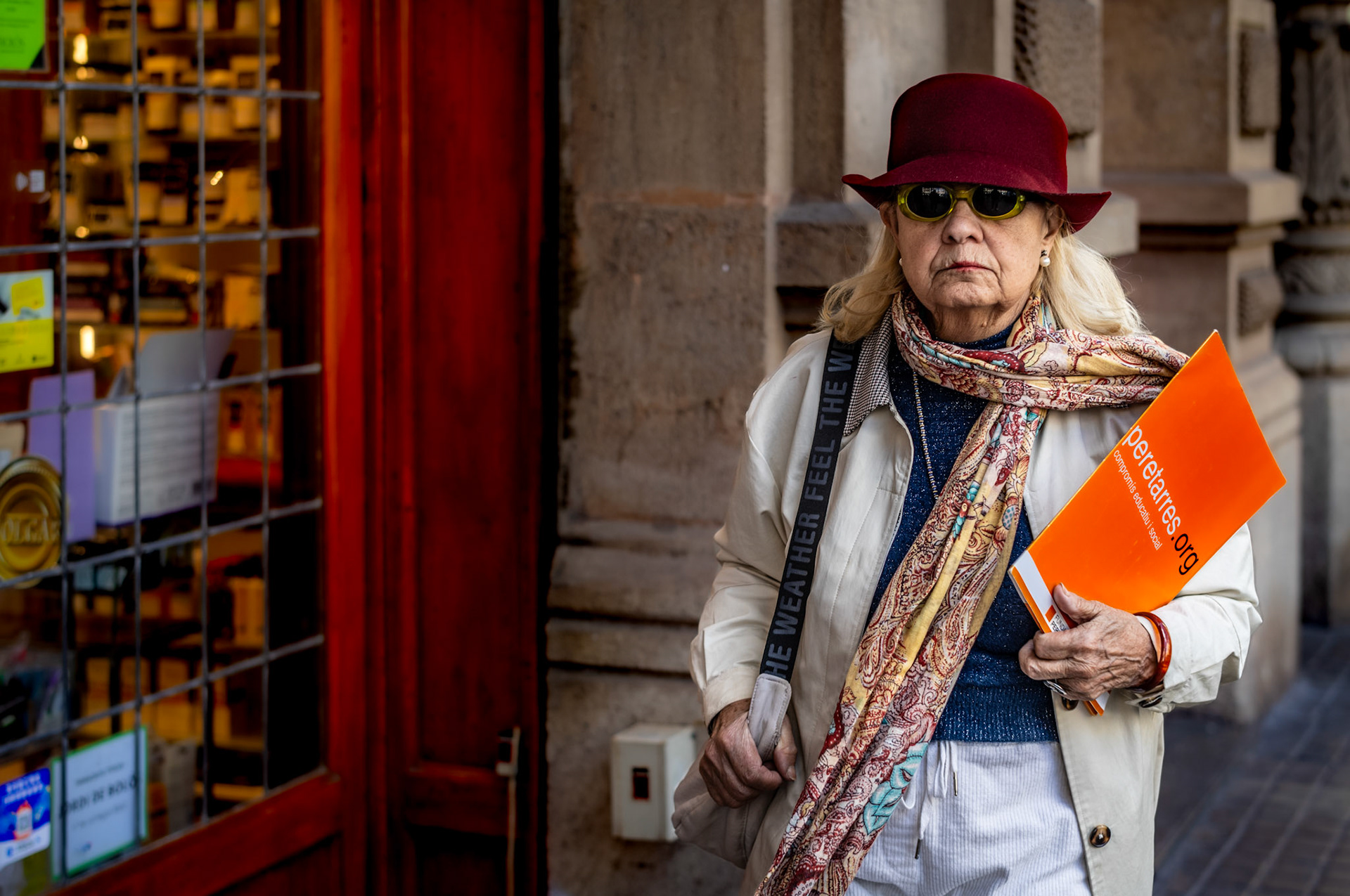 in the bustling heart of barcelona, amidst narrow lanes and vibrant storefronts, she stands out. her deep red hat, a shield against both sun and attention; the scarf, a swirl of stories; and that bright orange folder, a contrast against the urban tones, hinting at the world of business and movement. there's a determination in her stride, a hint of mystery behind those shaded glasses. captured in a fleeting moment, she becomes both observer and observed, a character that commands the frame, revealing the essence of life in the city.