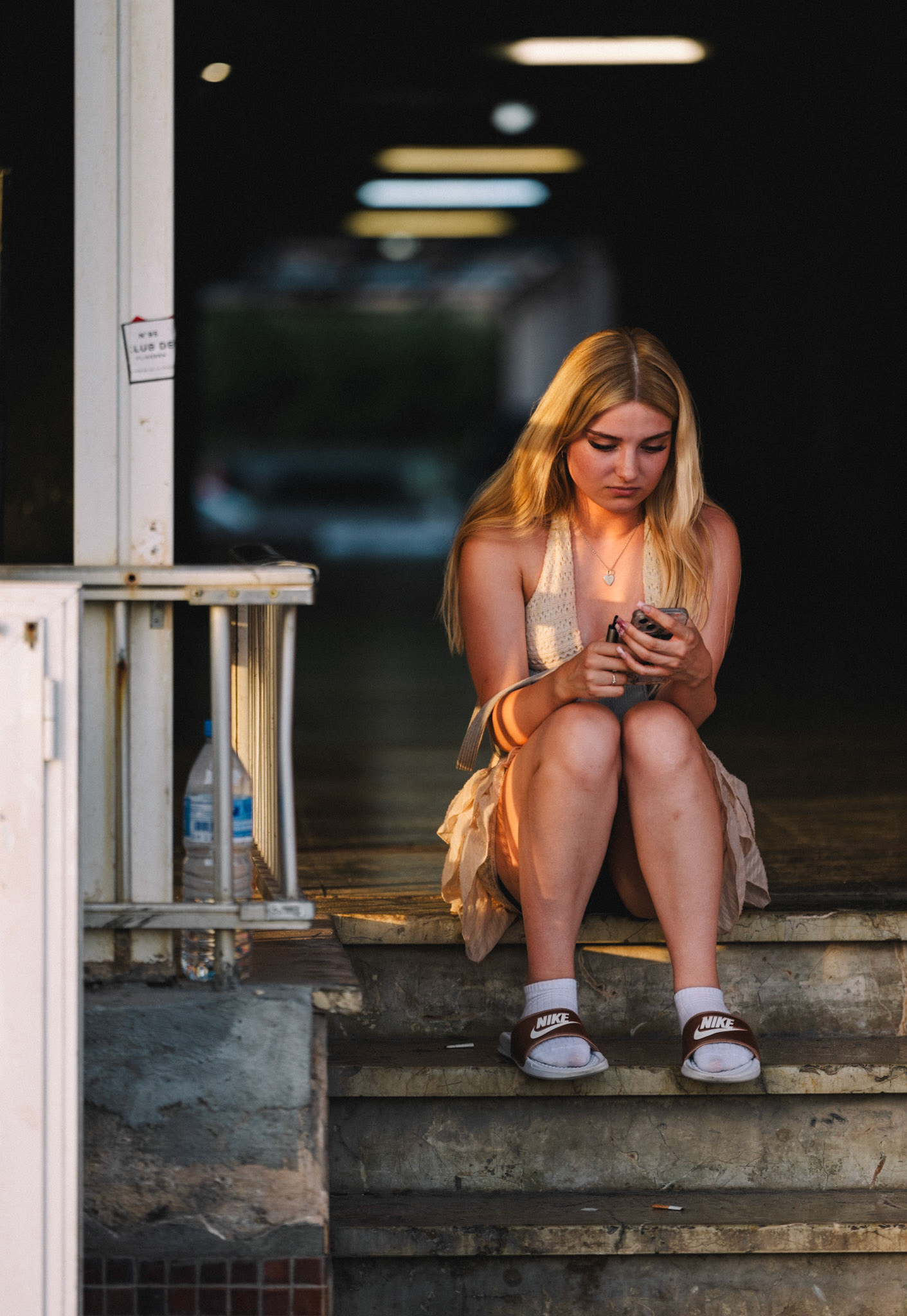 during a photowalk in the bustling el arenal of mallorca, i captured this quiet moment amidst the chaos. the soft, fading sunlight illuminates her, contrasting with the dark background, as she sits lost in her thoughts or perhaps her phone. it's a striking image of stillness in a place known for its energy. the subtle details, like the nike slides and the bottle, add a touch of reality to the scene.
