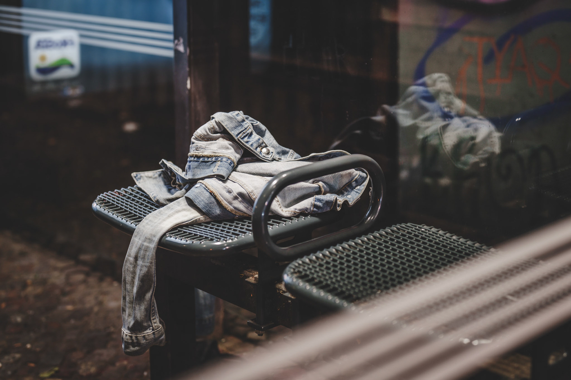 at the edge of the night, in the lonely glow of berlin's bus stop, an old denim jacket tells a story of abandonment. it lies forgotten, draped across the cold metal bench, illuminated only by the distant streetlights of the schlesisches tor. there's an unsettling quiet to this moment, as if the jacket's owner has vanished, leaving behind just this trace of their presence. the scene feels frozen, eerily still, a haunting reminder of lives passing through unnoticed, of the unspoken moments in the city that never sleeps.