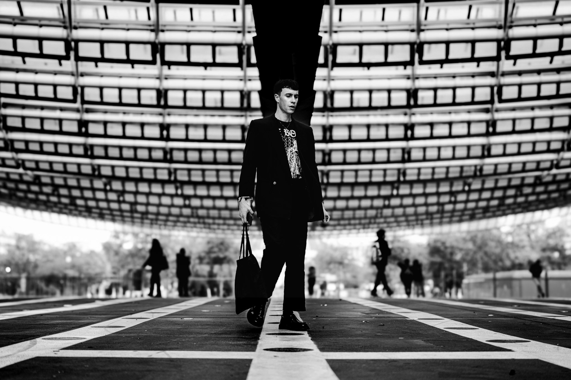 captured in the heart of paris, this image portrays a lone figure walking with quiet determination, as if moving between light and shadow. the architectural lines above stretch like a futuristic web, guiding the viewer's gaze directly to the man at the center. there's a sense of stillness and introspection in his steady steps, while the silhouettes in the background fade like distant memories, barely tangible. the black-and-white format emphasizes the geometry and rhythm of the lines, creating an atmosphere of detachment and contemplation. it's a silent, almost melancholic moment that reflects the pace of modern life, highlighting the contrast between the solitude of the individual and the busy world around him.