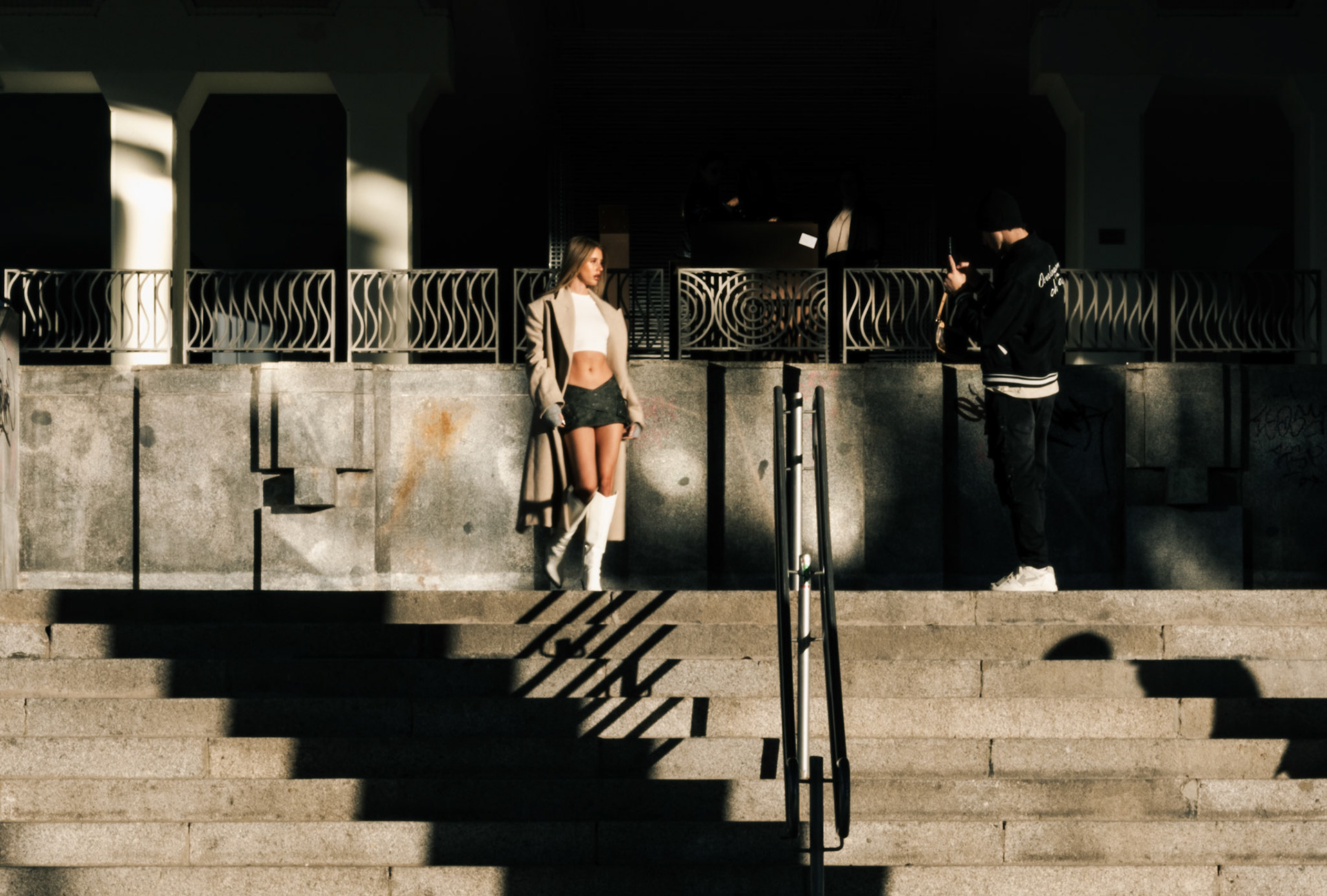 taken in the heart of madrid, this photo captures a young man photographing his stylish girlfriend against the backdrop of urban architecture. the warm glow of the setting sun highlights the subject, creating dramatic shadows and emphasizing the textures of the stone steps. the scene reflects the essence of modern street photography, where everyday moments are immortalized through the lens of a smartphone. it’s a glimpse into the world of the instagram generation, where everyone is a potential influencer, and every moment is a chance to create art.