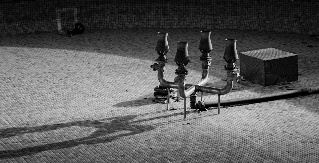 in the crisp embrace of winter, the fountain's dormant nozzles stand like a sculptor's interpretation of a candelabra, an unintentional work of art born from functionality. the city has turned its bustling symphony down to a whisper, allowing the fountain's quietude to take center stage. with its water stilled, the nozzles cast intricate shadows upon the herringbone canvas below, a pattern that speaks to the order amidst the stillness. the lone wheel, perhaps a forgotten toy or a discarded relic, adds a touch of whimsy to the scene, a silent player in this off-season tableau. as the winter light drapes over this urban still life, the photograph captures the beauty of pause, the elegance of the offbeat, and the serene anticipation of spring's eventual return.