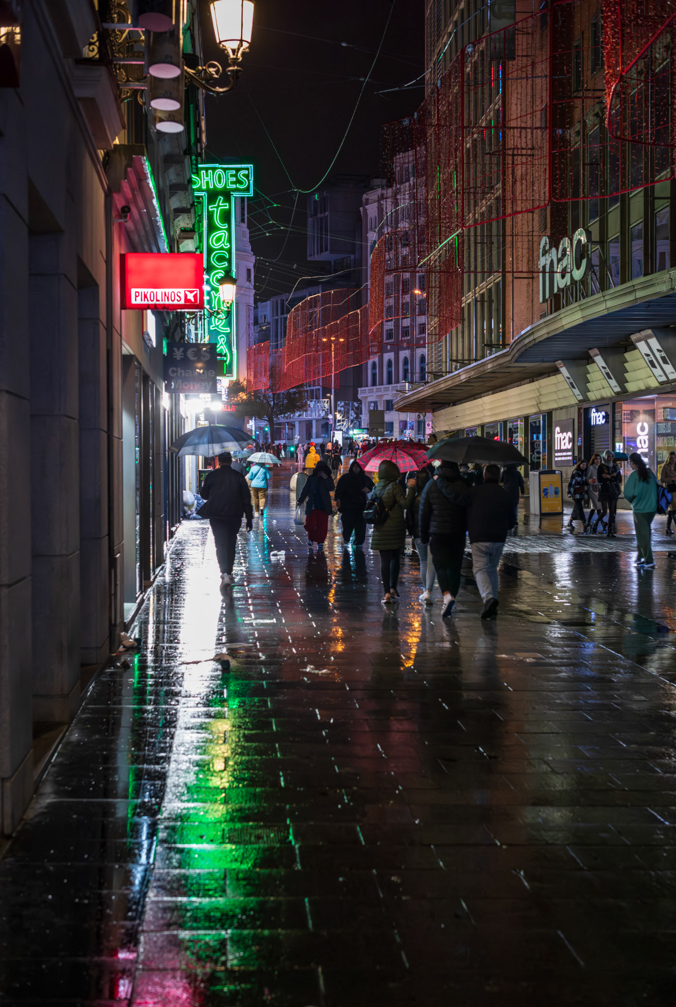captured in madrid during a rainy night in wintertime, the city streets come alive with the reflections of neon lights on the wet pavement. people with umbrellas weave through the vibrant bustle, creating a dynamic atmosphere that only a nocturnal downpour can evoke. the energy of the city is palpable, even in the rain.