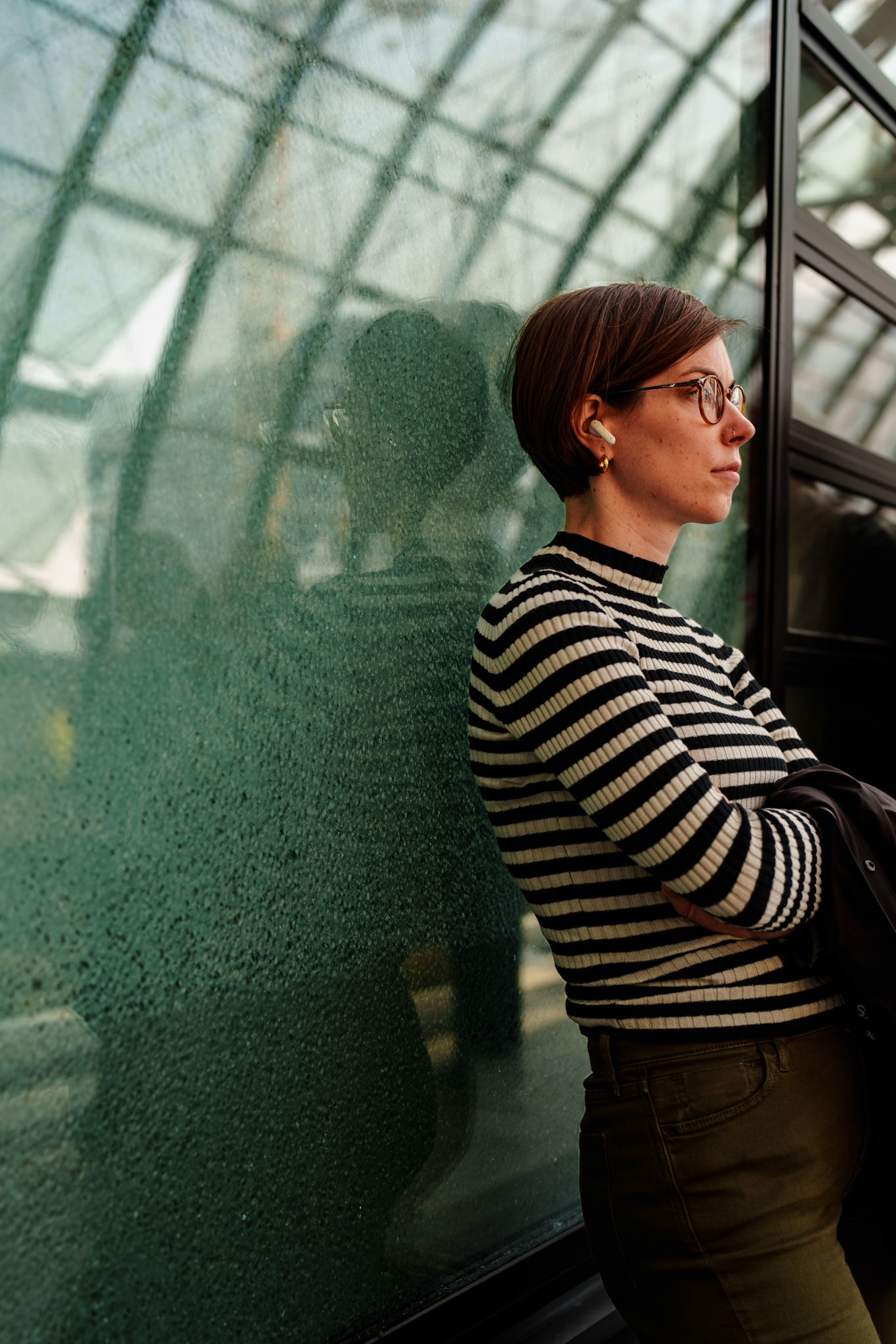 at the heart of berlin's central station, she stands with poise, her reflection blending with the rain-splattered glass. there's a contrast between the structured lines of her striped sweater and the fluid shapes forming behind her on the window. absorbed in her thoughts, she waits, a calm presence amidst the city’s constant motion, both mirrored and isolated in the architectural expanse.