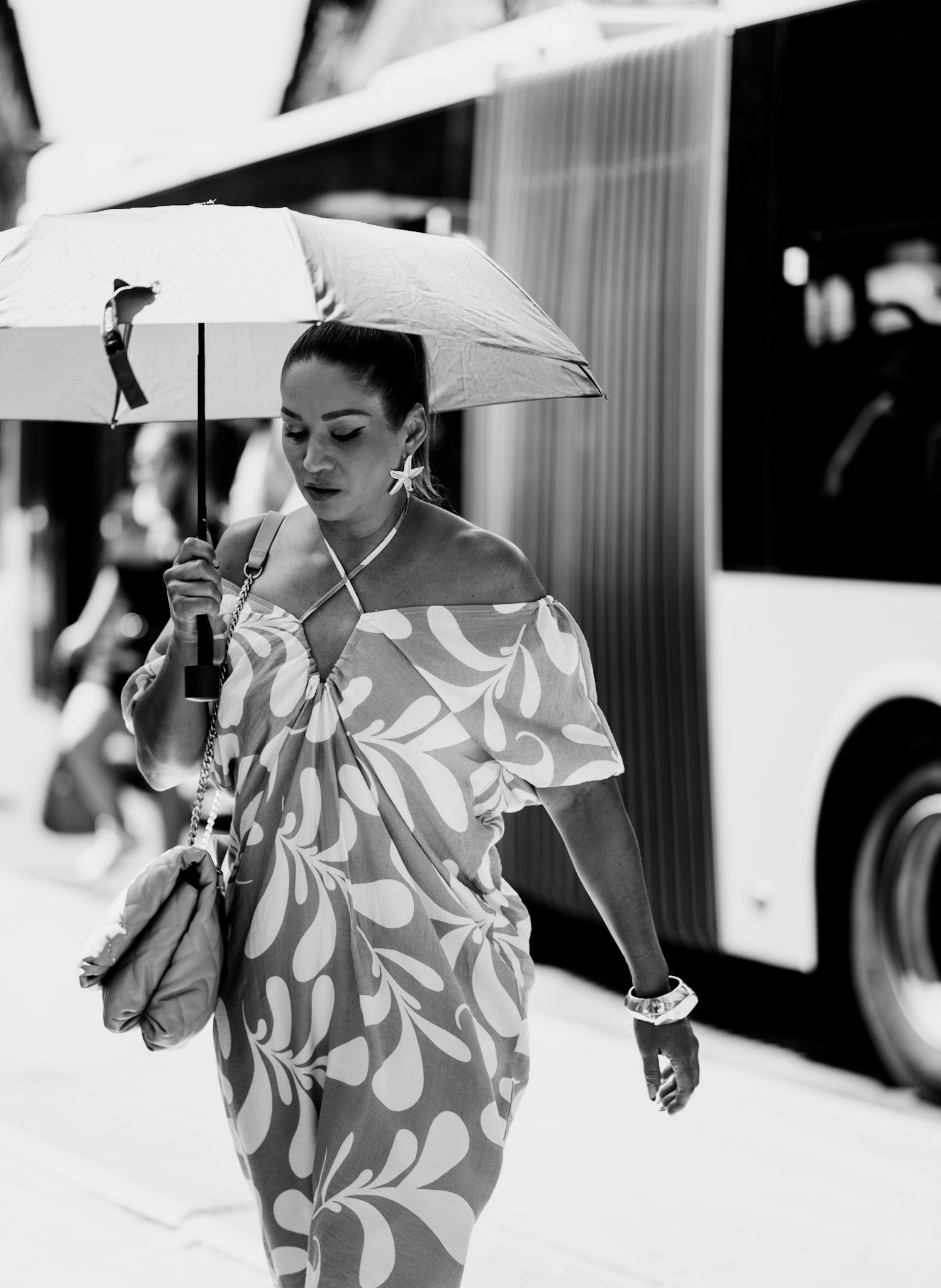 in the sweltering heat of a 38-degree summer evening in palma, the sight of a woman stepping off the bus with a rain umbrella catches the eye—an unexpected contrast against the sun's relentless blaze. as she moves with poise and a quiet confidence, her ensemble radiates elegance. the patterned dress, off-shoulder and flowing, echoes the curves of her every step, and the accessories—from the statement earrings to the polished bag—complete a look that is both refined and effortlessly chic. amidst the bustle, the heat, and the chaos, she embodies a moment of calm, where fashion meets function, and style transcends the ordinary.